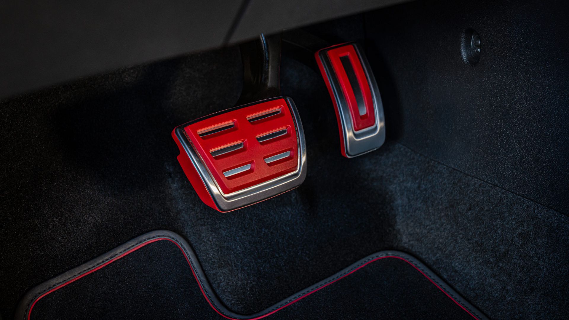 Detailný záber na pedále auta s čiernym koberčekom. Pedále majú červené a strieborné akcenty. Brzdový pedál je vľavo a pedál akcelerátora vpravo.