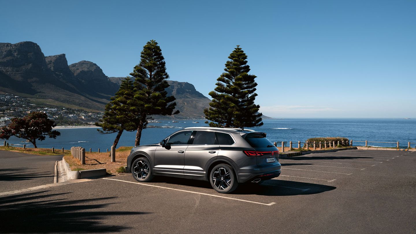 VW Touareg Elegance zaparkovaný na slnku, pohľad zboku, za ním je vidieť more a pobrežie.
