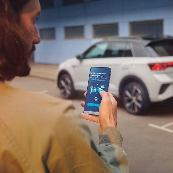 VW T-Roc R-Line v bielej farbe pred bytovým domom modrej farby, vozidlo je rozostrené. V popredí stojí muž so smartfónom, na ktorom aktivuje aplikáciu Volkswagen.