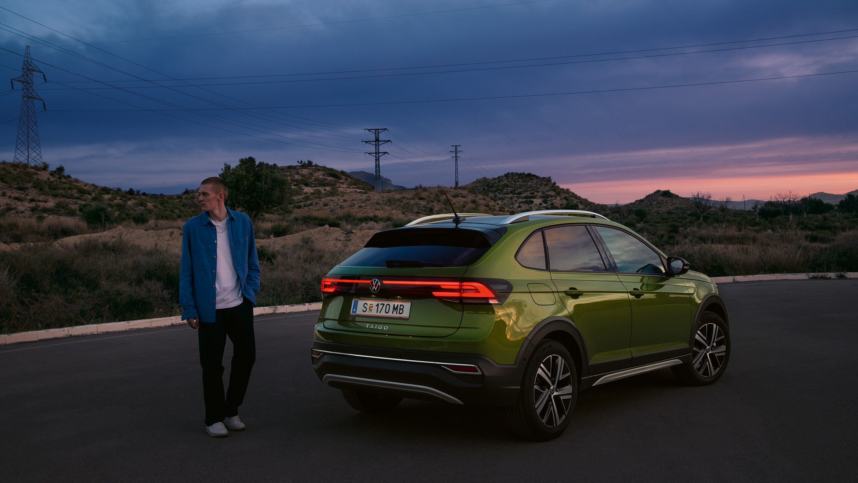VW Taigo Style v zelenej farbe je zaparkovaný na parkovisku na horách. Muž stojí vedľa neho. V pozadí vidieť menšie kopce. Celú scénu dotvára večerná atmosféra pri západe slnka.