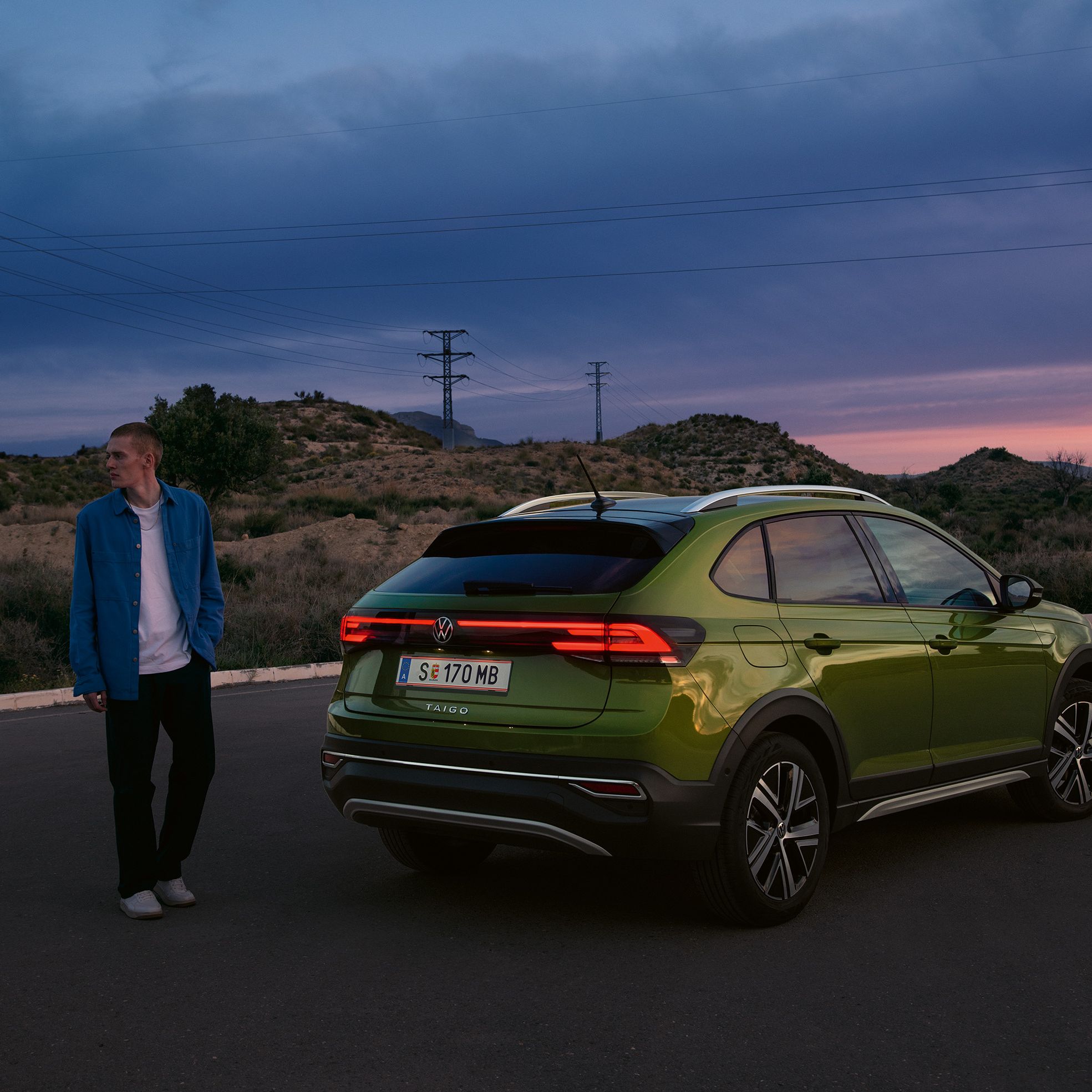 VW Taigo Style v zelenej farbe je zaparkovaný na parkovisku na horách. Muž stojí vedľa neho. V pozadí vidieť menšie kopce. Celú scénu dotvára večerná atmosféra pri západe slnka.