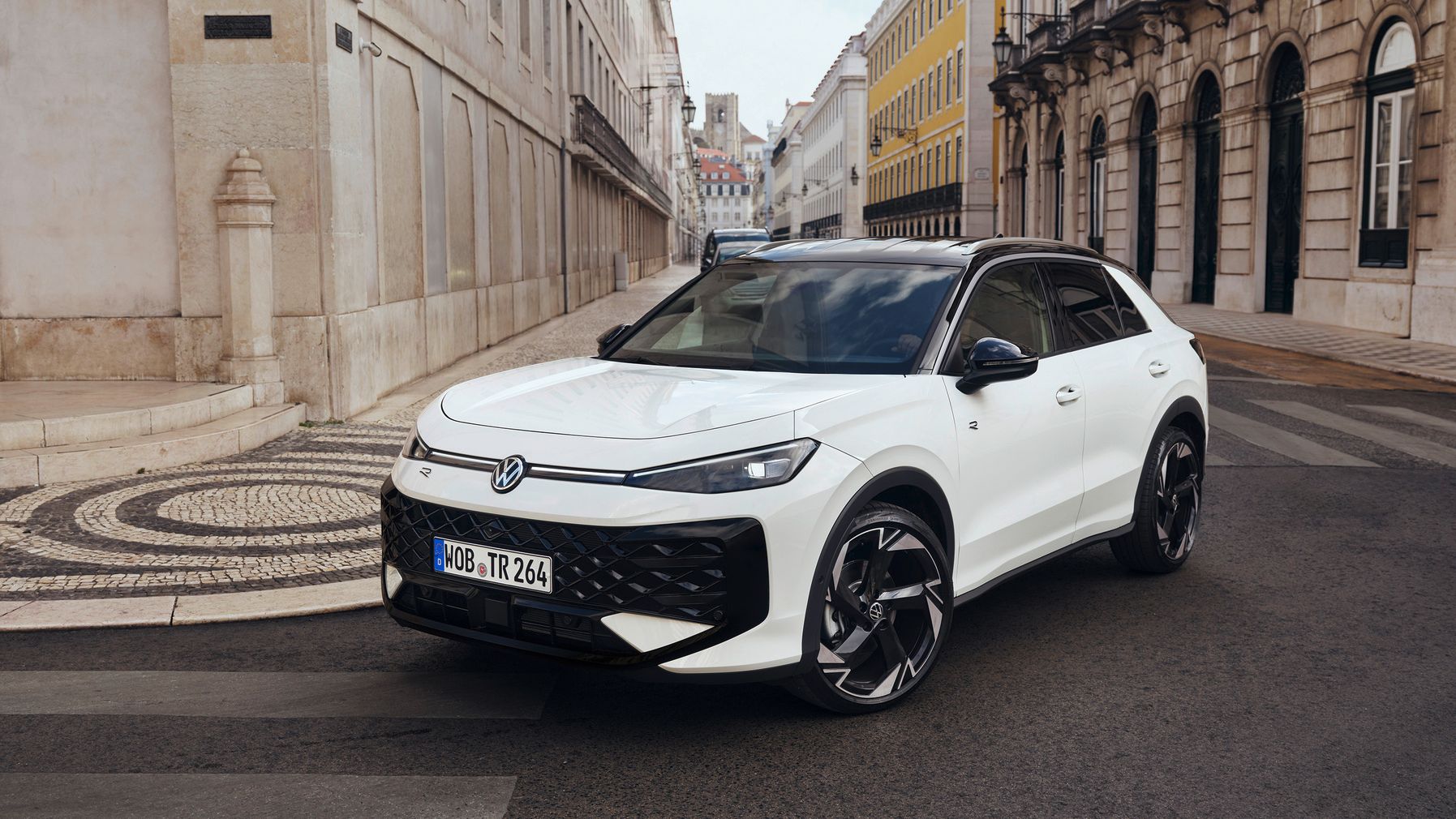 Biely Volkswagen T-Roc je zaparkovaný na ulici s historickými budovami. Má športový dizajn s čiernymi doplnkami a modernými líniami. Okolo sú dlažobné kamene a ozdobné lampy.