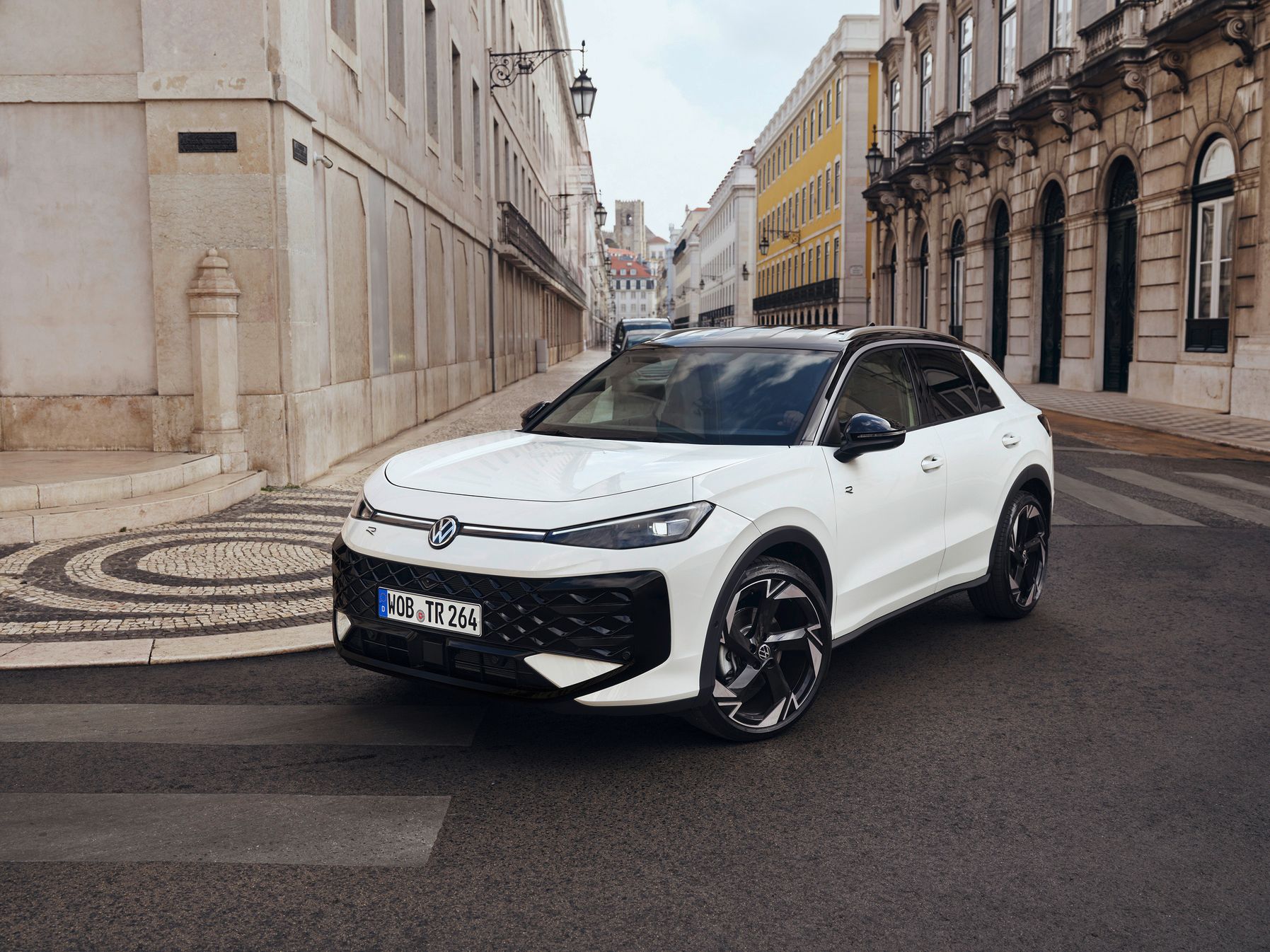 Biely Volkswagen T-Roc je zaparkovaný na ulici s historickými budovami. Má športový dizajn s čiernymi doplnkami a modernými líniami. Okolo sú dlažobné kamene a ozdobné lampy.