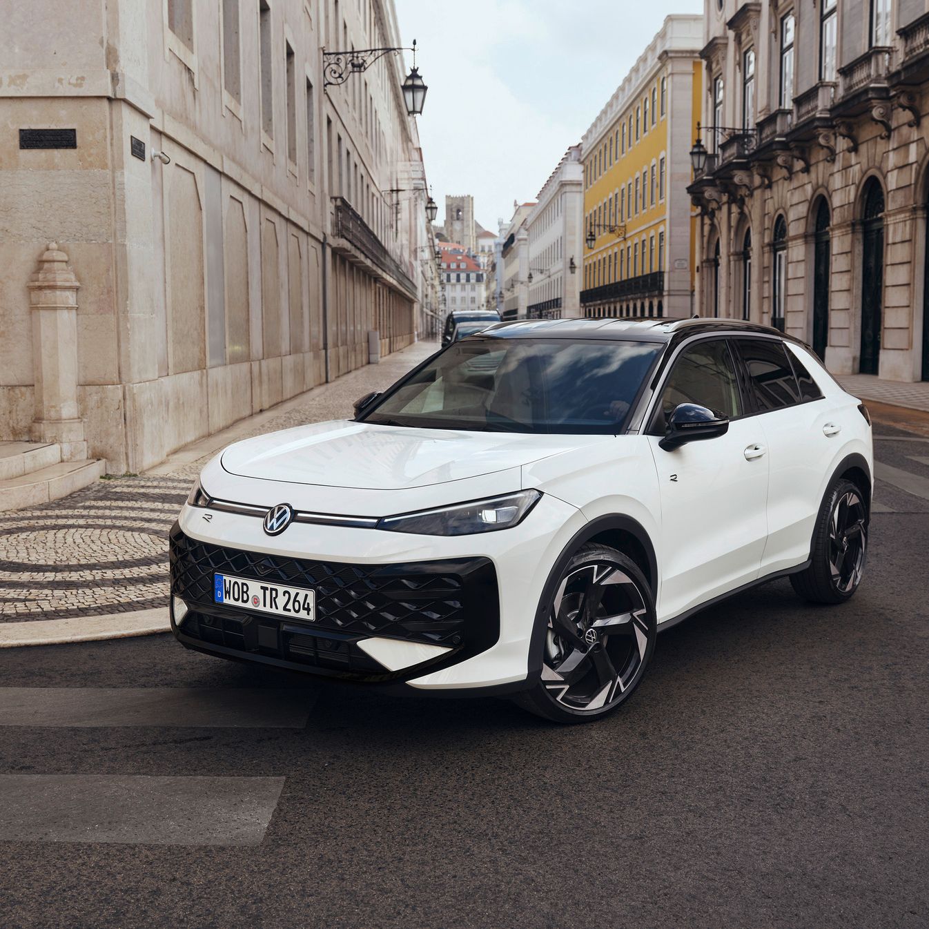 Biely Volkswagen T-Roc je zaparkovaný na ulici s historickými budovami. Má športový dizajn s čiernymi doplnkami a modernými líniami. Okolo sú dlažobné kamene a ozdobné lampy.