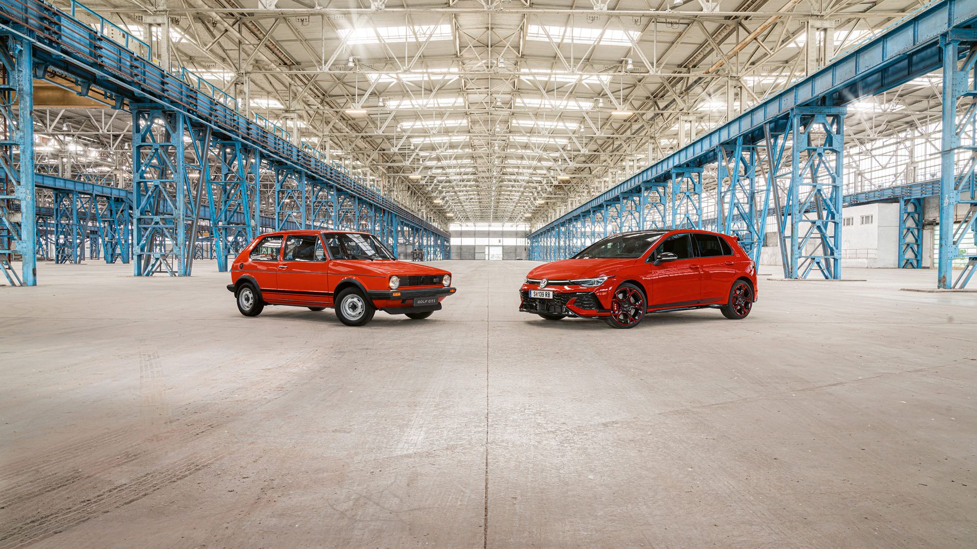 Dva červené modely Golf GTI, prvý a najnovší model, stoja vo veľkej priemyselnej hale s modrou oceľovou konštrukciou a vysokou, svetlou strechou.