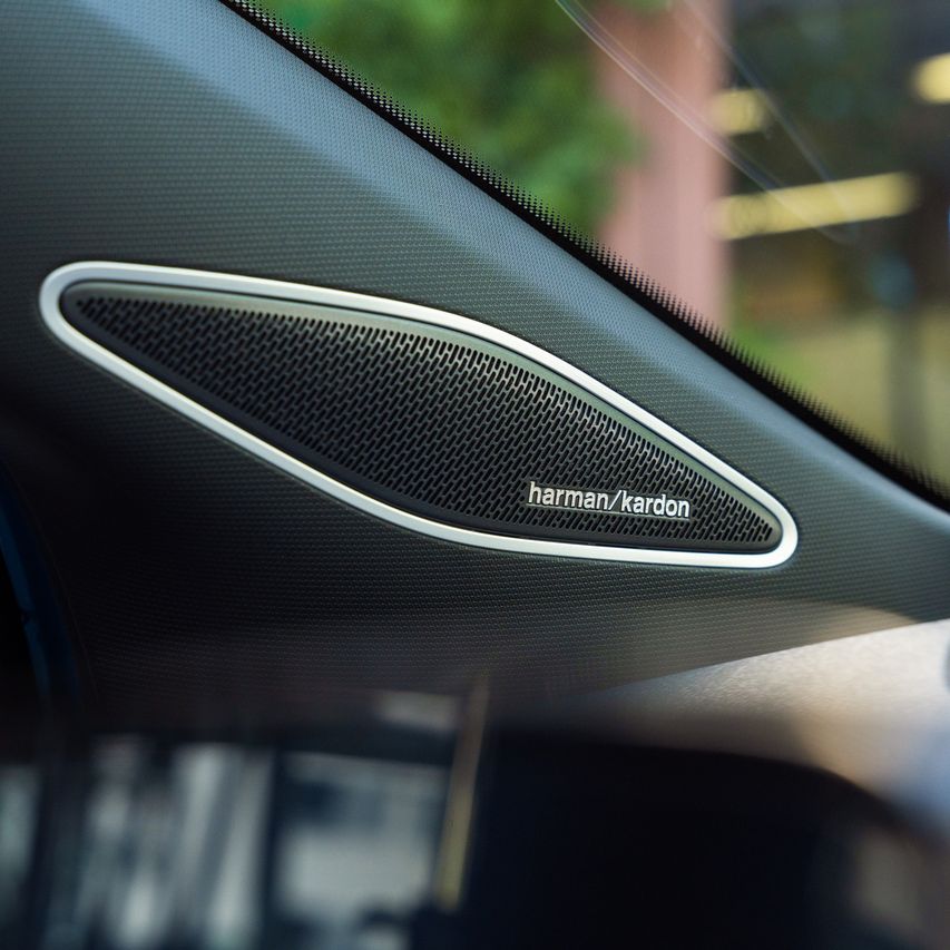 Pohľad na reproduktor Harman/Kardon integrovaný v A-stĺpiku. Mimo detailu je okno auta, cez ktoré čiastočne vidieť vonkajšie prostredie.