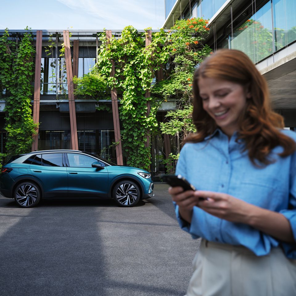 Moderné SUV zaparkované pred budovou pokrytou zeleňou. Žena s rozmazanou tvárou drží telefón a stojí v popredí.