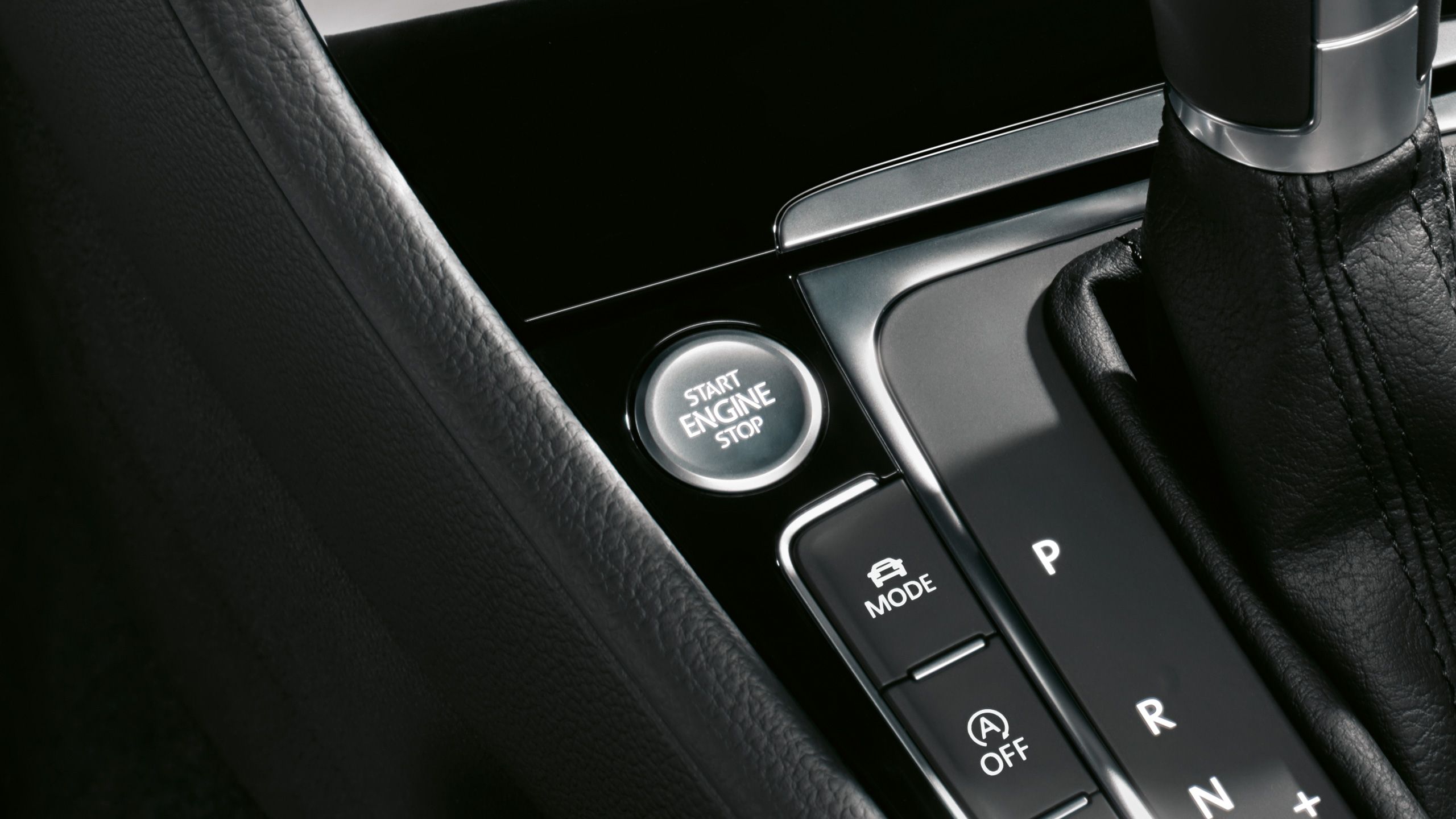 Tlačidlo na naštartovanie a vypnutie motora v modernom vozidle. Pod tlačidlom sú ďalšie ovládacie prvky automatickej prevodovky. Panel je z čierneho lesklého materiálu.