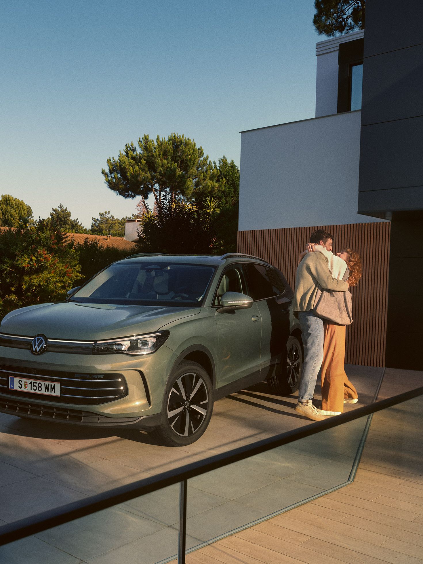 VW Tiguan parkuje pri modernej vile. Vedľa vozidla stoja v objatí muž a žena. Okolo pozemku je veľa zelene.