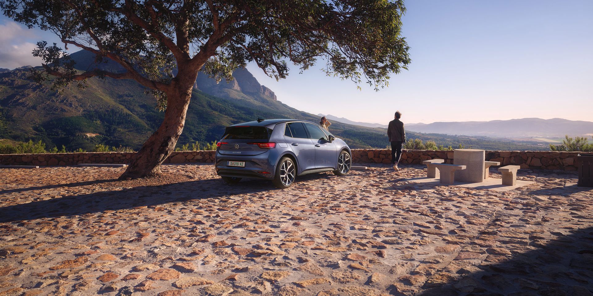 Na parkovisku v horách stojí modrý VW ID.3 Neo, žena a muž si užívajú výhľad.
