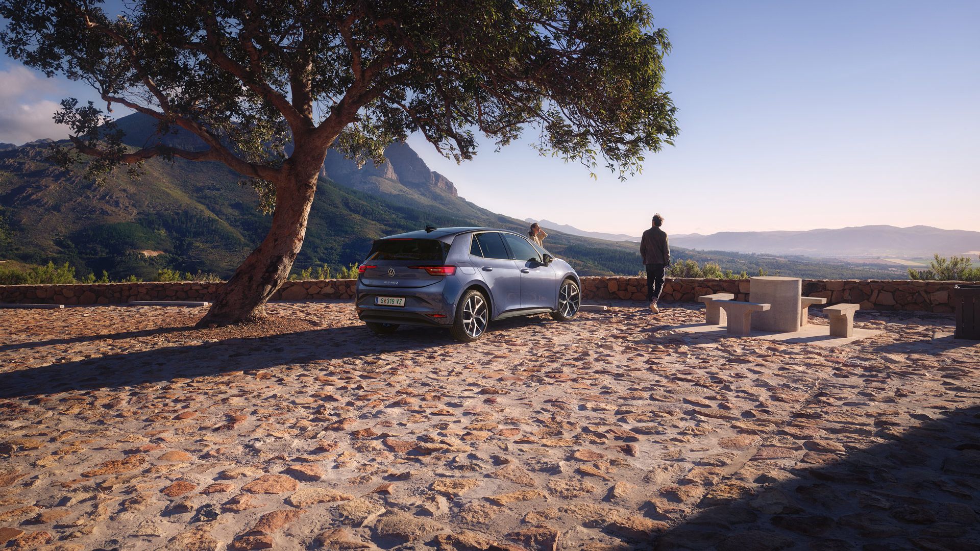 Na parkovisku v horách stojí modrý VW ID.3 Neo, žena a muž si užívajú výhľad.