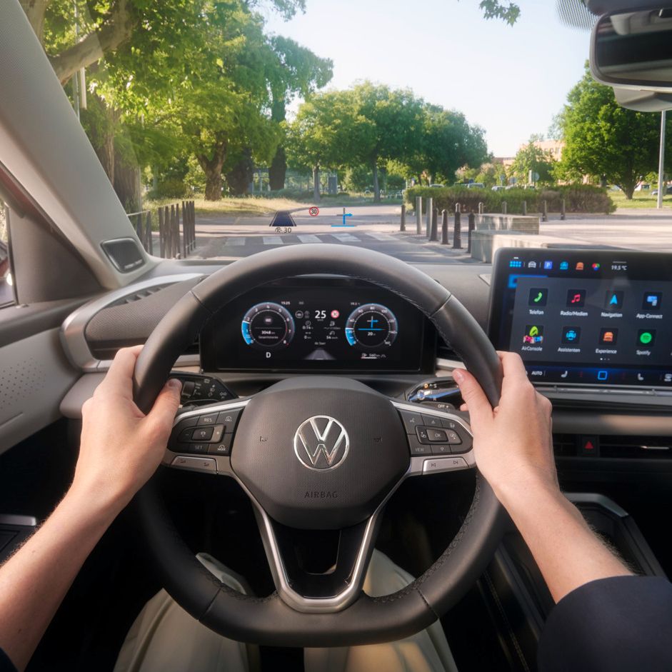 Pohľad na prístrojový panel a displej MMI v VW T-Roc Style s detailom na head-up displej na čelnom skle. Cez čelné sklo vidieť cestu, vpravo modernú, verejnú budovu a vľavo mestský park..