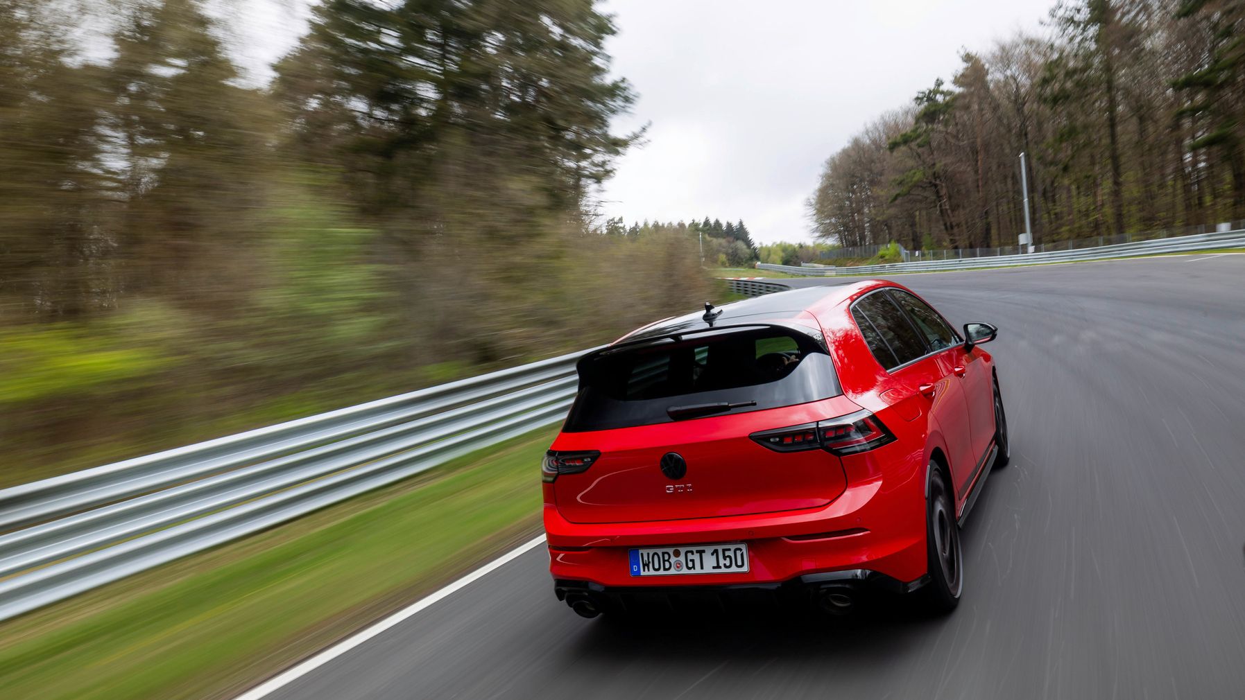 Červené športové auto Volkswagen GTI jazdí po asfaltovej ceste obklopenej stromami. Zadná strana auta je viditeľná so značkou ŠPZ. Automobil dynamicky prechádza zákrutou.