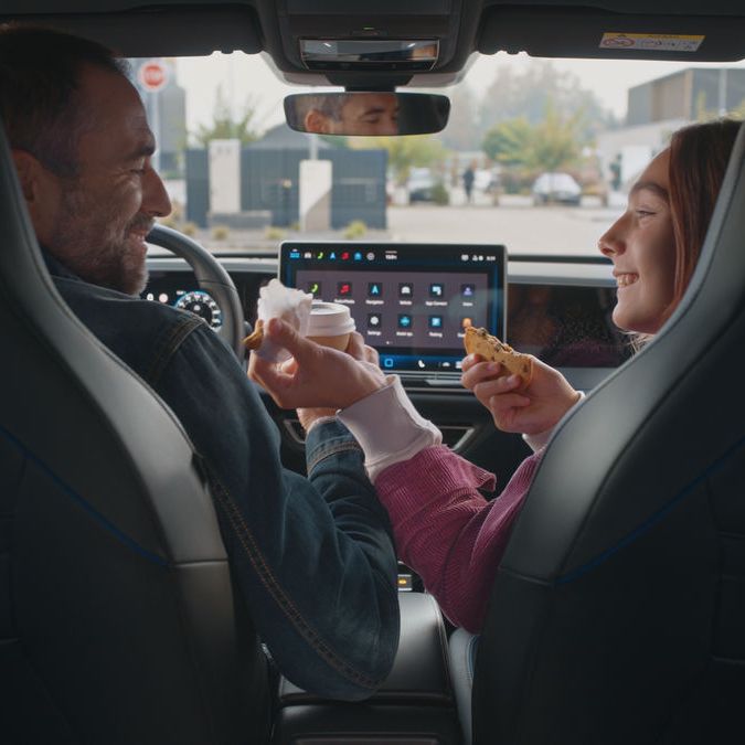 Dvaja ľudia sedia v aute. Jeden z nich podáva druhému kúsok jedla. Na stredovej obrazovke auta je zobrazený infotainment systém.