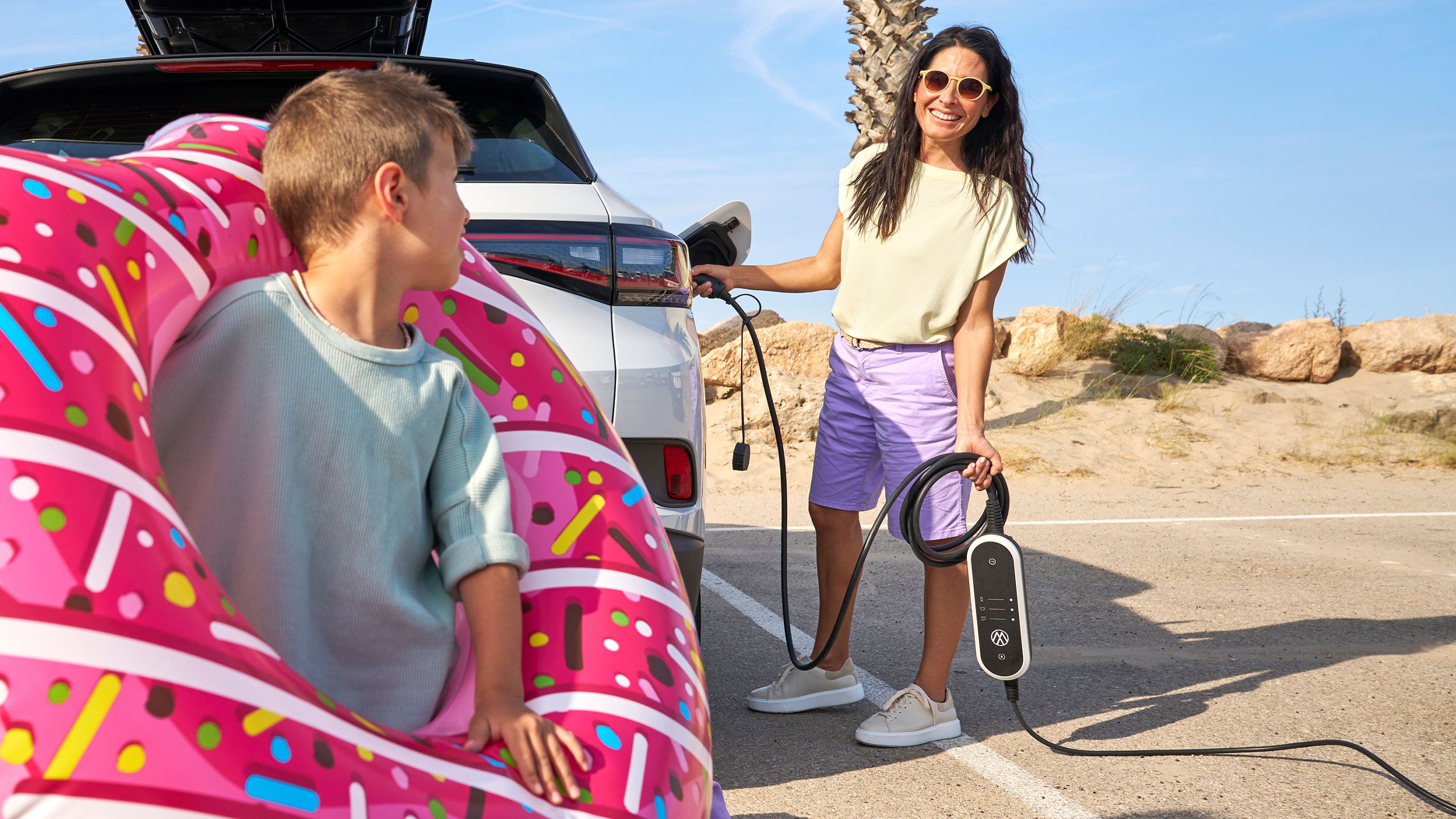Dieťa s plaveckým kruhom, žena nabíja model VW ID s cestovnou nabíjačkou ID Charger Basic od VW príslušenstva  v pozadí.