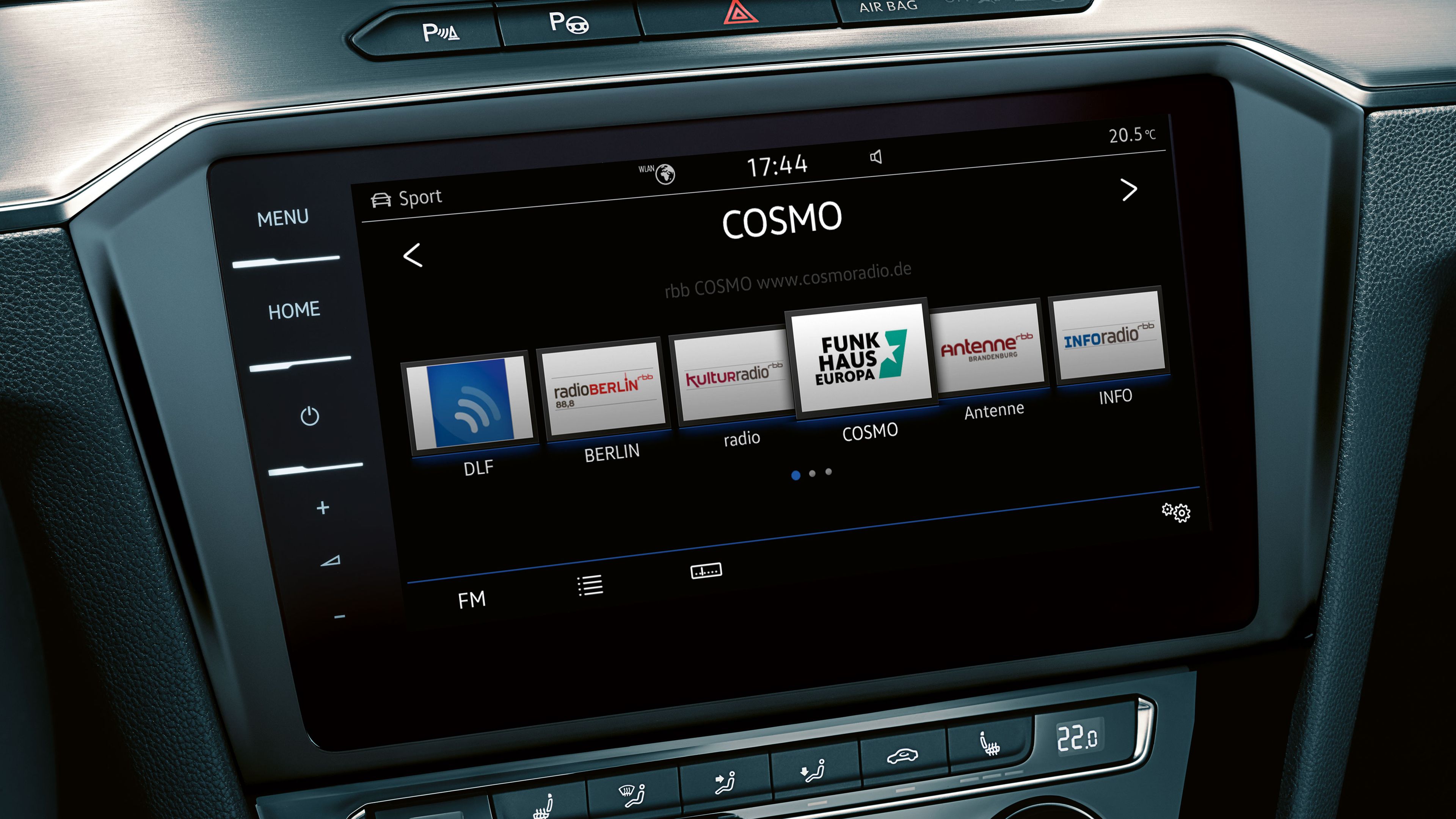 Na obrázku je dotykový displej systému infotainment, na ktorom je zvolené rádio COSMO. Okolo displeja sú tlačidlá na ovládanie rádia, klimatizácie a ďalších funkcií vozidla.