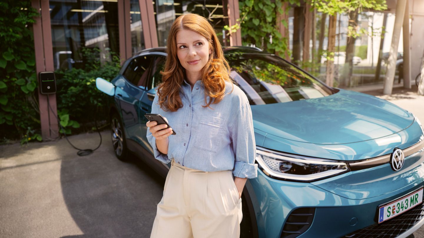 Žena so smartfónom v ruke stojí pri modrom VW ID.4, ktorý je pripojený k nabíjacej stanici.