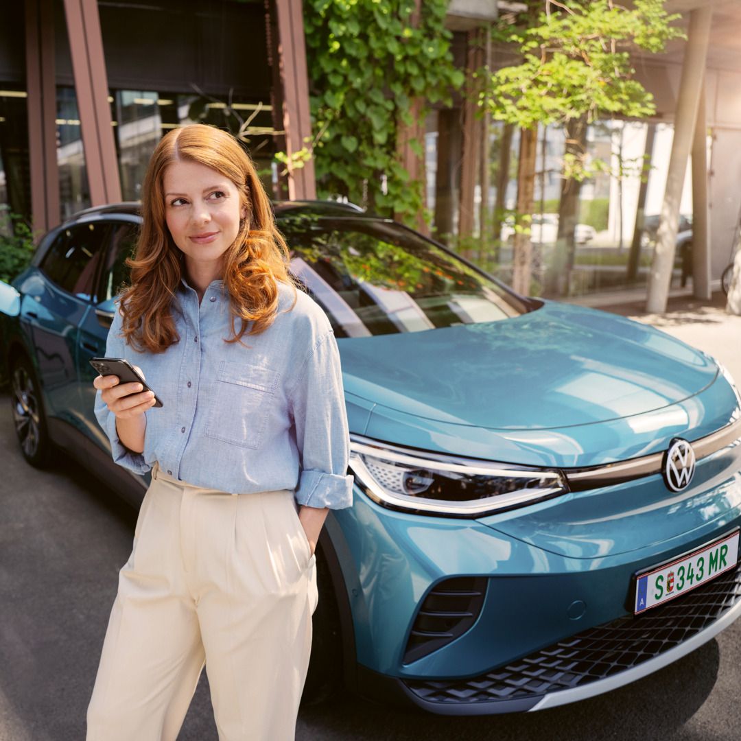 Žena so smartfónom v ruke stojí pri modrom VW ID.4, ktorý je pripojený k nabíjacej stanici.