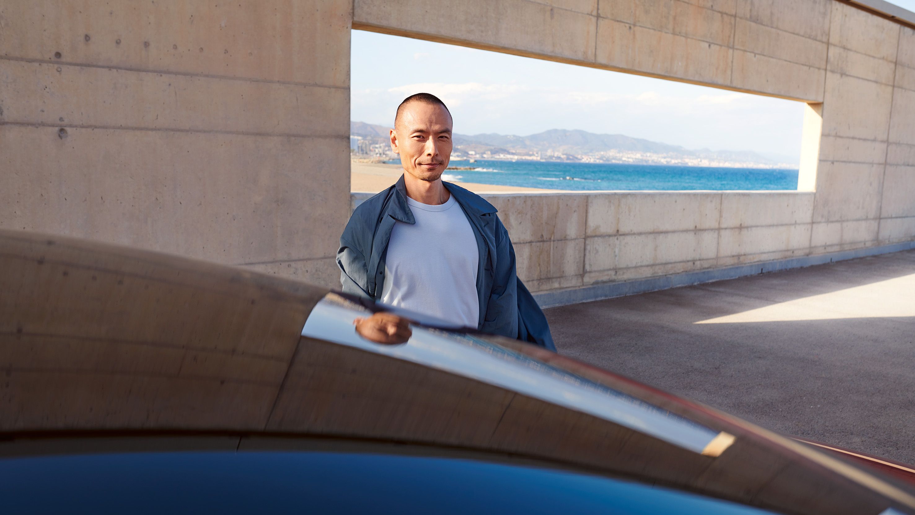 Muž stojí pri vozidle na parkovisku pri betónove stene s veľkým otvorom  s výhľadom na more, pláž a mesto.