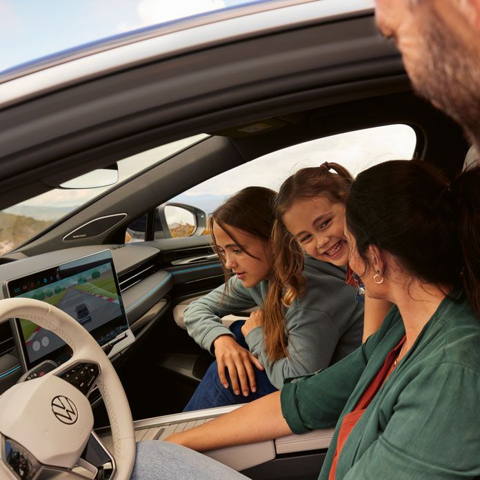 Rodina testuje hry v aplikácii vo vozidle VW ID.7. Žena sedí na sedadle vodiča, jedno dievča vedľa nej a jedno sa nakláňa dopredu zo zadných sedadiel. Muž pozerá dovnútra cez  otvorené dvere vodiča.