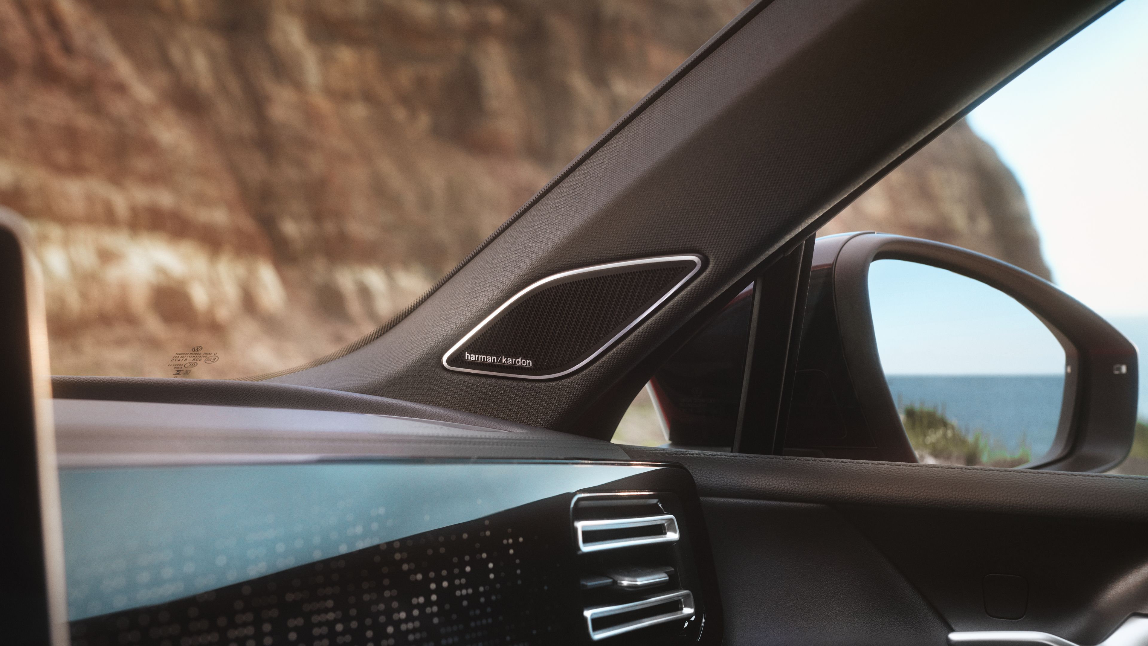 Detail interiéru VW Tiguan so zameraním na reproduktor inteegrovaný do A-stĺpika. Čiastočne vidieť palubnú dosku s ambientným ovládaním, vonkajšie spätné zrkadlo a cez okná výhľad na útes.