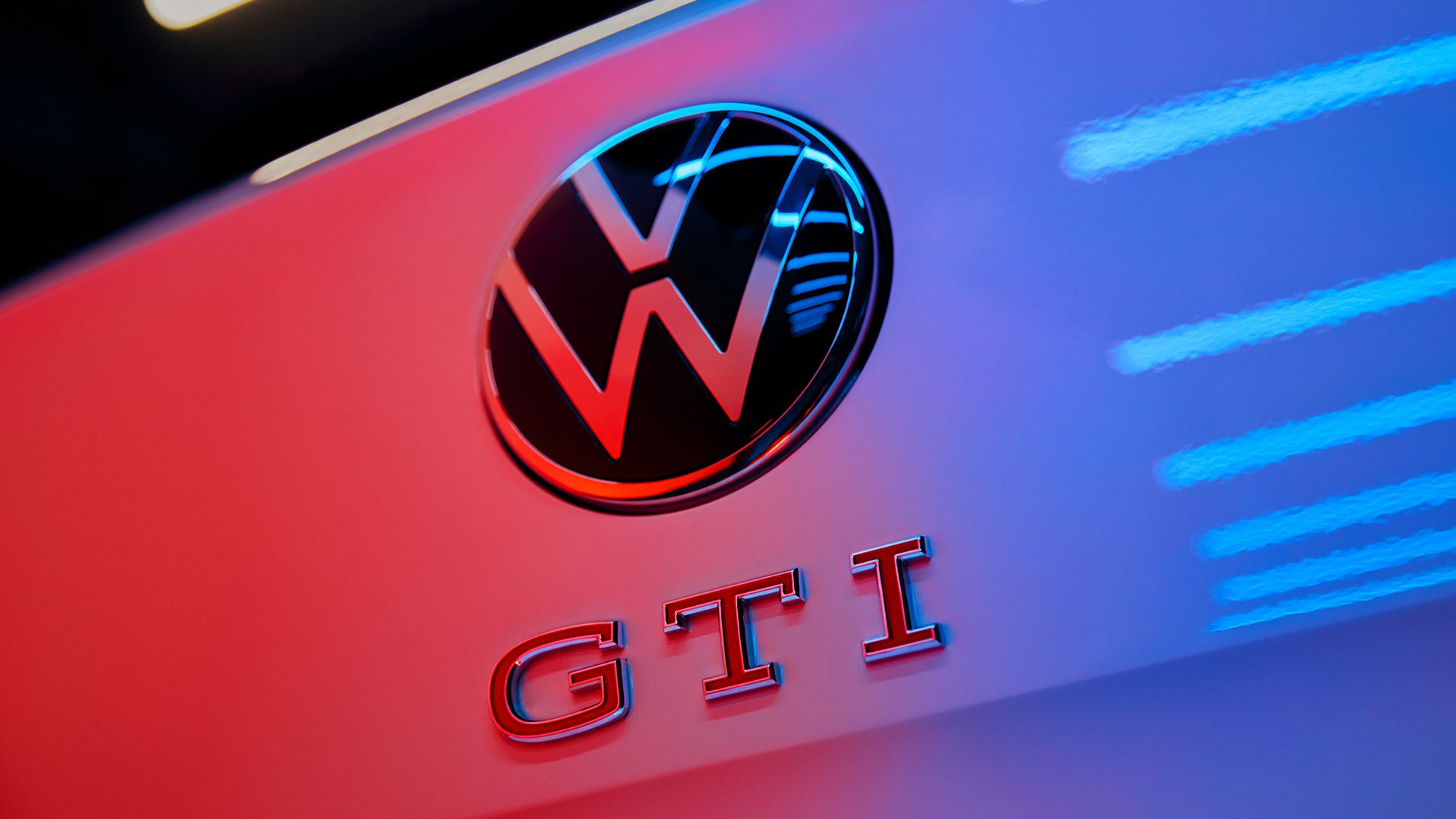 Logo Volkswagen GTI na zadných dverách osvetlené modro červeným svetlom. V dolnej časti je nápis GTI. Okolo loga a nápisu sú rozmazané svetelné pruhy.