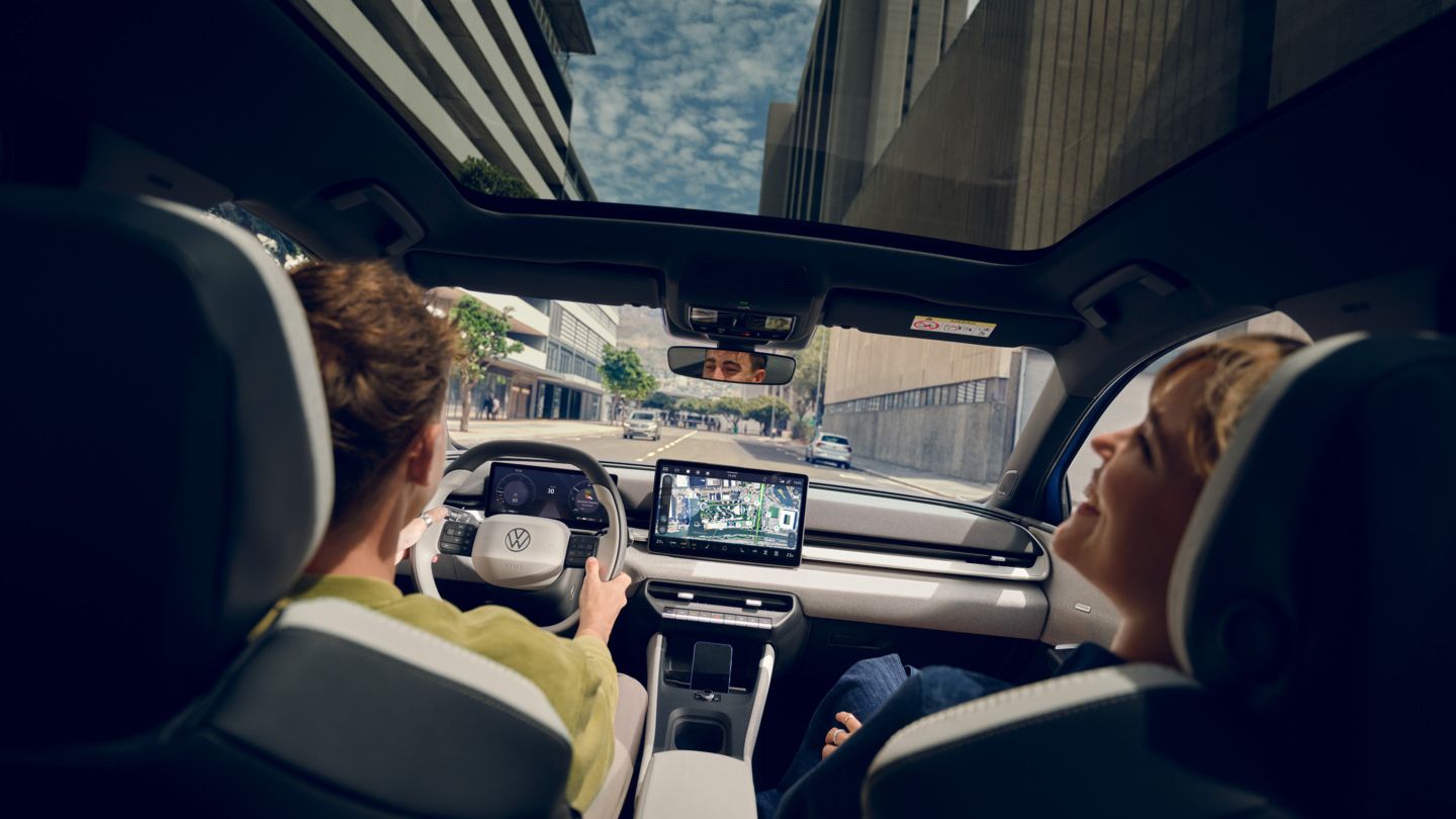 Muž riadi VW ID. Polo Style, na sedadle spolujazdca sedí usmievajúca sa žena. Cez panoramatickú strechu sú vidieť výškové budovy.