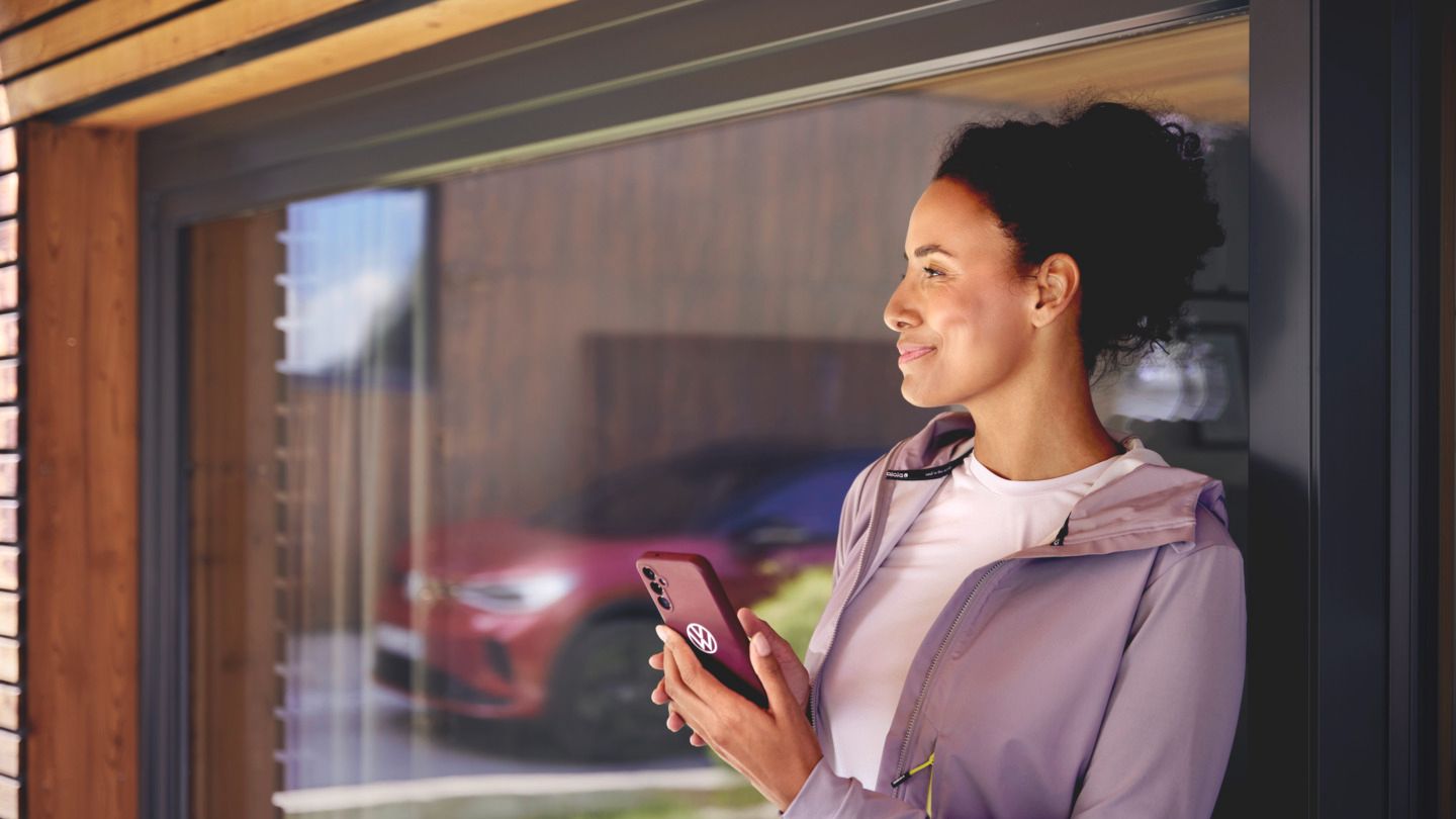 Žena stojí pri veľkom sklenenom okne domu. V ruke drží smartfón a v odraze skla je vidieť červený ID.4 GTX.