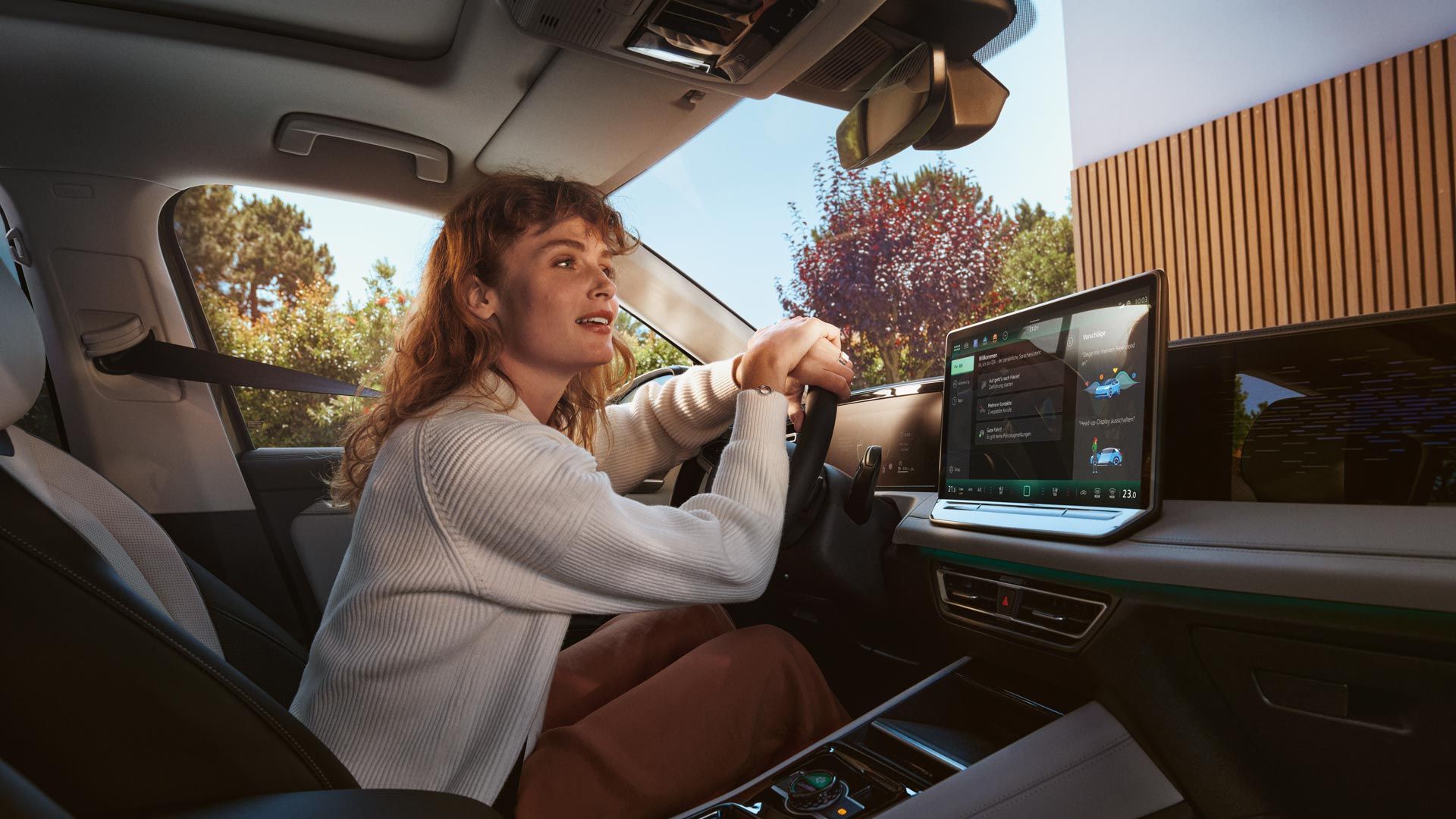 Žena sedí na sedadle vodiča, ruky má na volante a nakláňa sa k čelnému sklu. V aute je veľký displej systému infotainment na palubnej doske s funkciami. Von je slnečné počasie a vidieť stromy a budovu pred vozidlom.