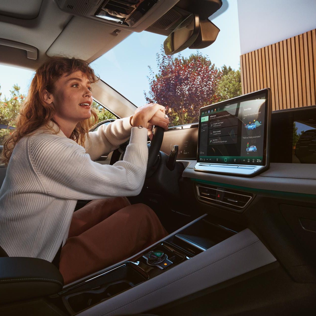 Žena sedí na sedadle vodiča, ruky má na volante a nakláňa sa k čelnému sklu. V aute je veľký displej systému infotainment na palubnej doske s funkciami. Von je slnečné počasie a vidieť stromy a budovu pred vozidlom.