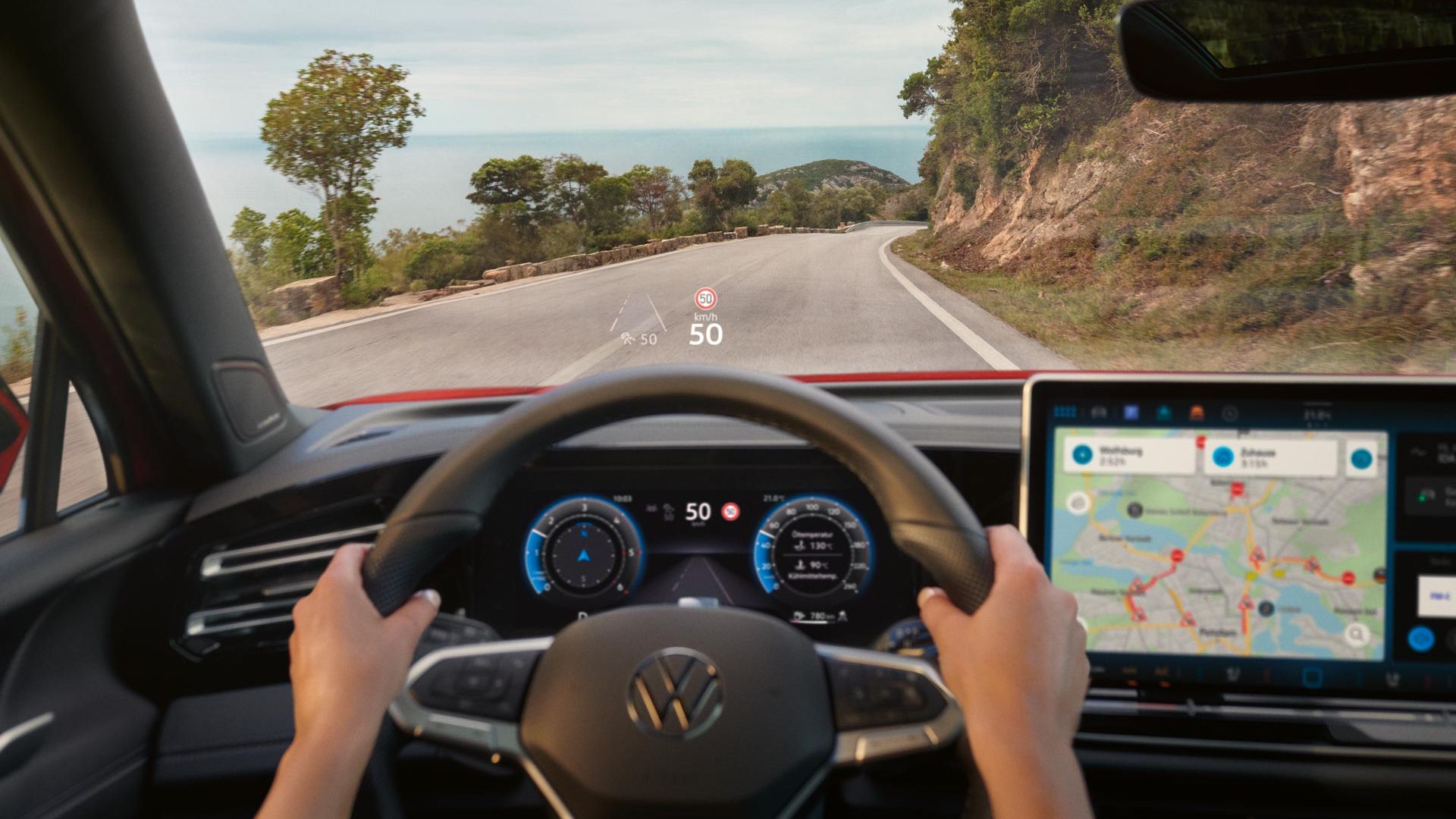 Pohľad do interiéru auta na volant a digitálny prístrojový panel, displej systému infotainment s navigačnou mapou počas jazdy po horskej ceste. Vodič drží volant. Cez čelné sklo vidieť cestu je ohraničená stromami a skalnou stenou.