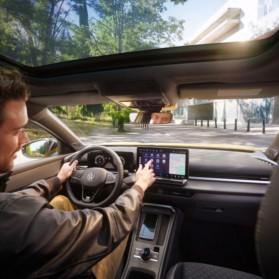 Muž s hnedými vlasmi a čiernou bundou sedí na sedadle vodiča vo VW T-Roc a ovláda systém infotainment, pričom druhou rukou drží volant. Vozidlo stojí na parkovacom mieste vedľa cesty, v pozadí je vpravo vidieť modernú budovu a vľavo park..