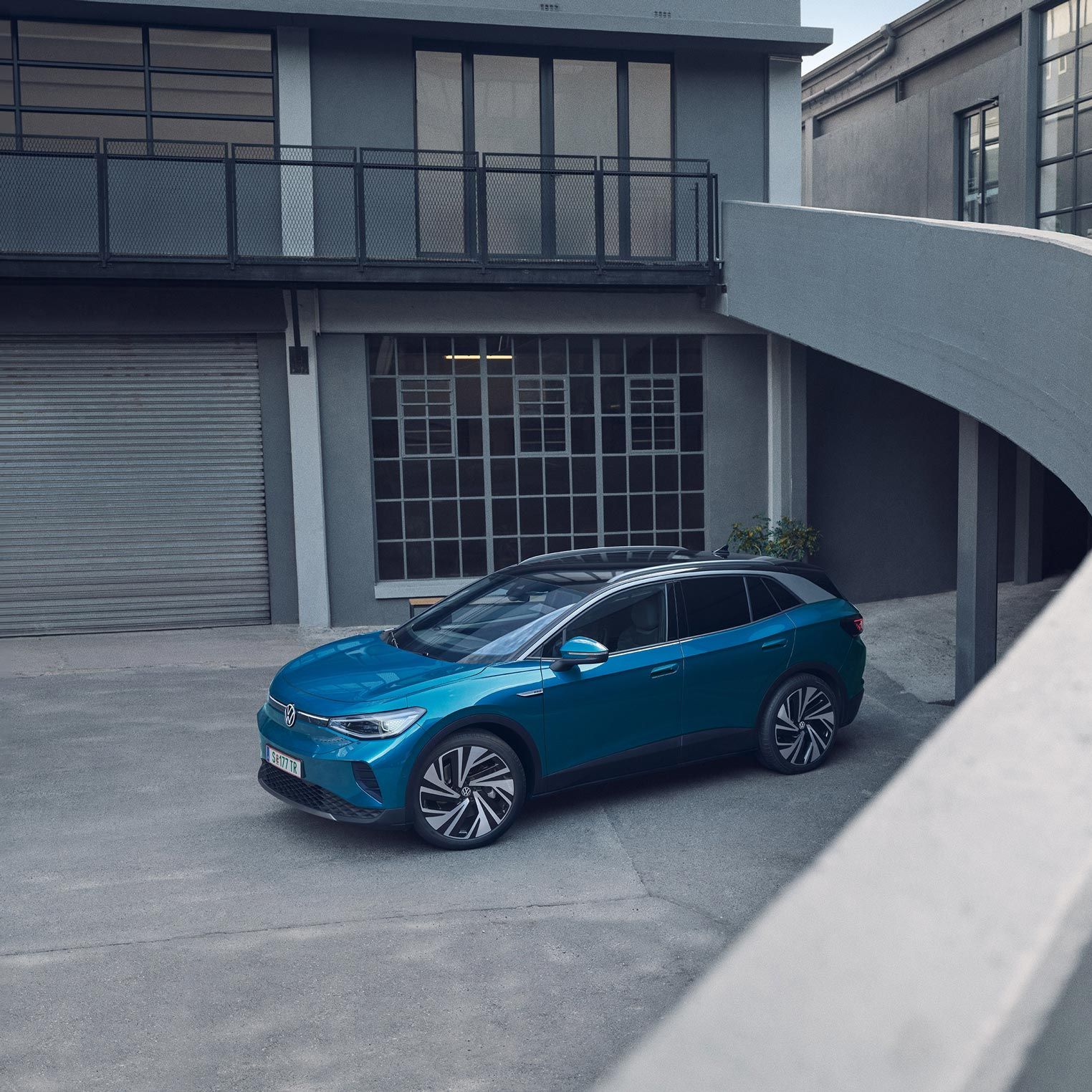 Modré SUV je zaparkované na betónovej ploche vedľa moderných budov. Auto má štýlový dizajn. Scéna je pokojná bez prítomnosti ľudí.