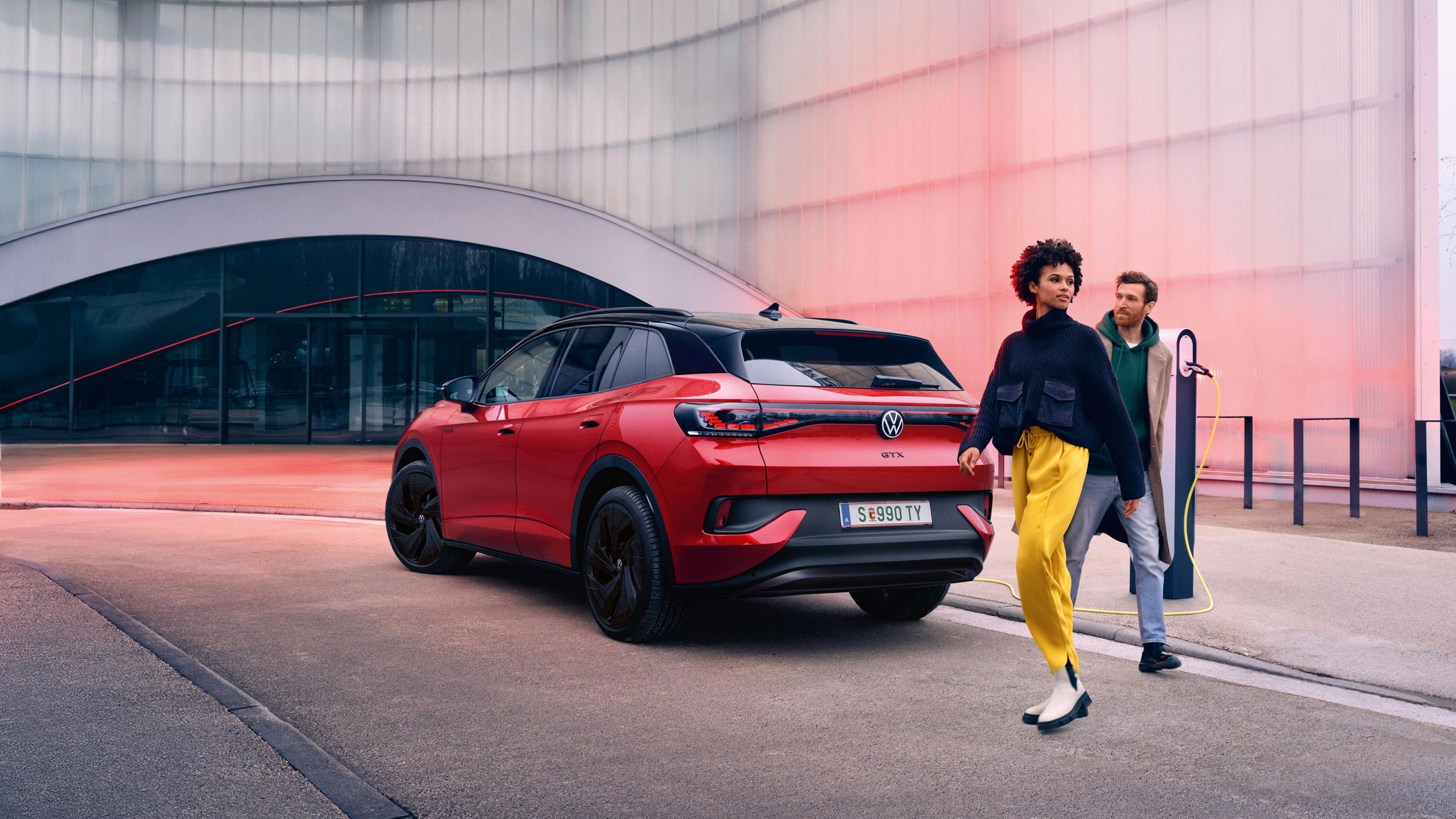 Pohľad na červený VW ID.4 GTX zozadu a zboku. Nabíja sa pri ceste, žena a muž prechádzajú okolo. Stojí pri  modernej budove s priehľadnou fasádou.