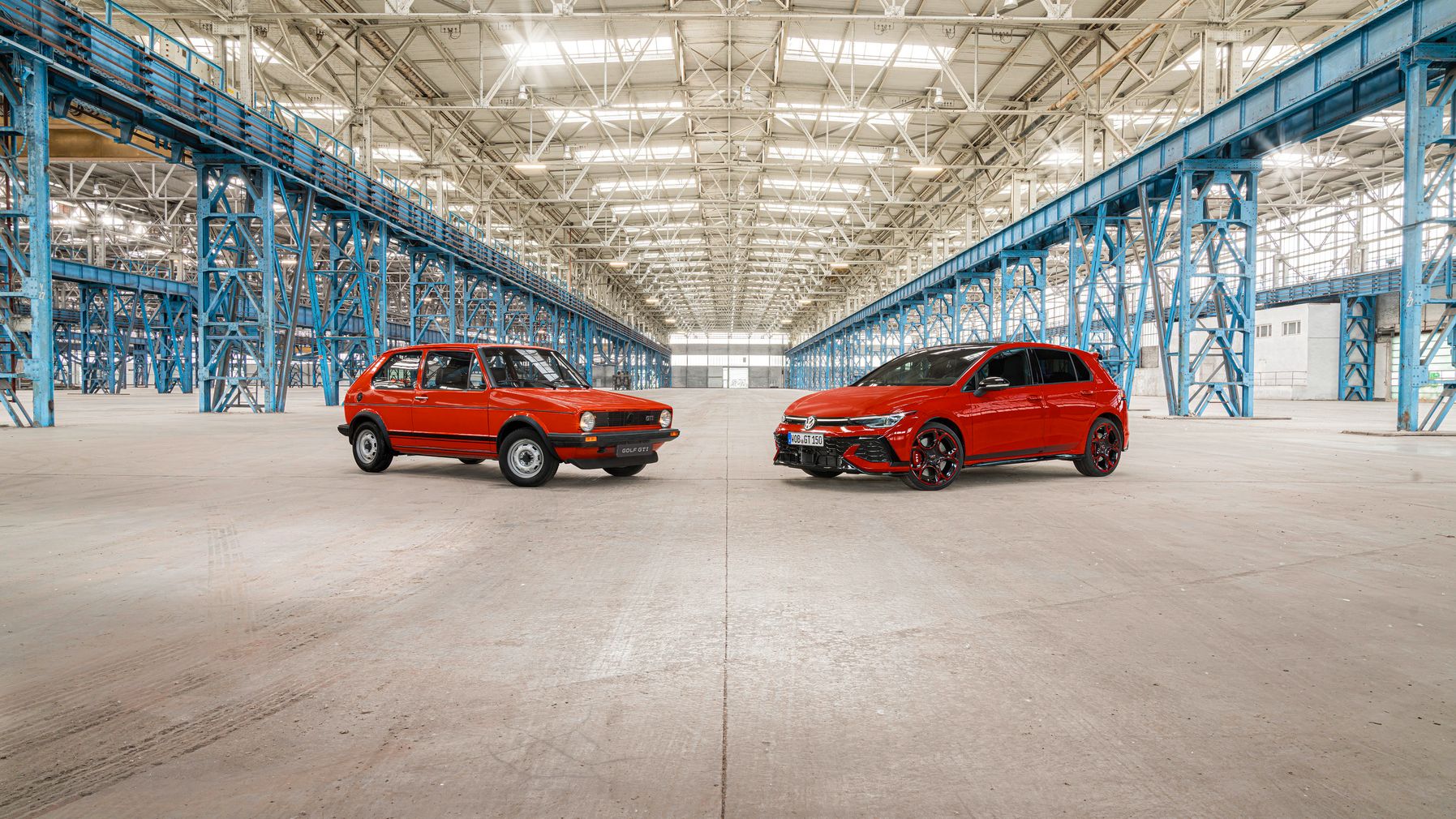 Dve červené vozidlá, jedno staršie a jedno moderné, sú zaparkované vedľa seba v prázdnej veľkej hale. Hala má kovovú konštrukciu a je veľmi priestranná.