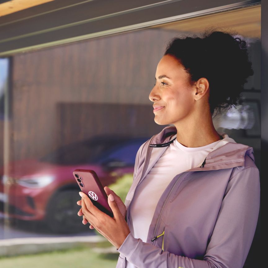 Žena stojí pri veľkom sklenenom okne domu. V ruke drží smartfón a v odraze skla je vidieť červený ID.4 GTX.