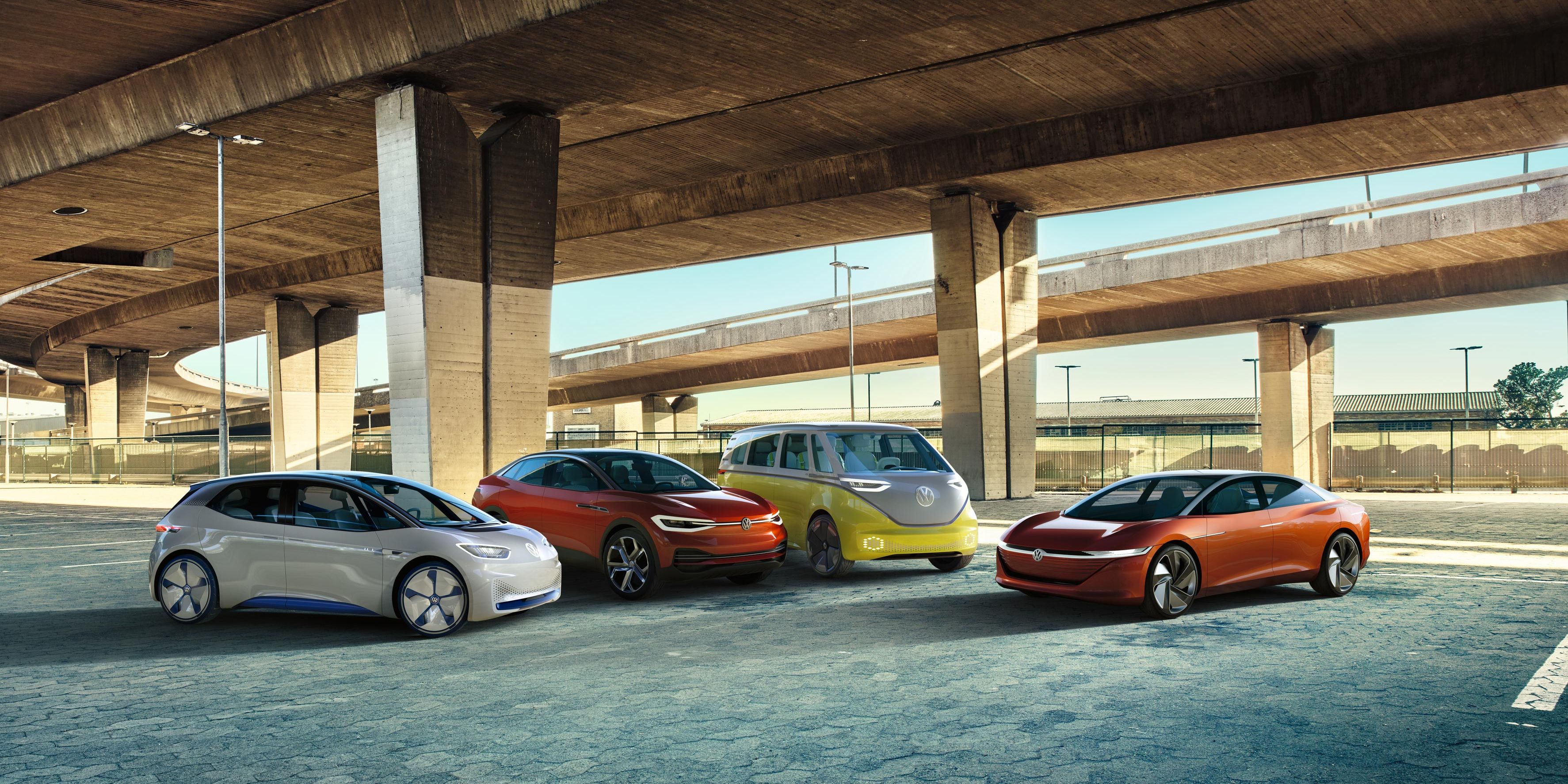 Pod mostom je zaparkovaných niekoľko moderných elektrických vozidiel Volkswagen rôznych farieb. Miesto pôsobí prázdne a má priemyselný nádych.