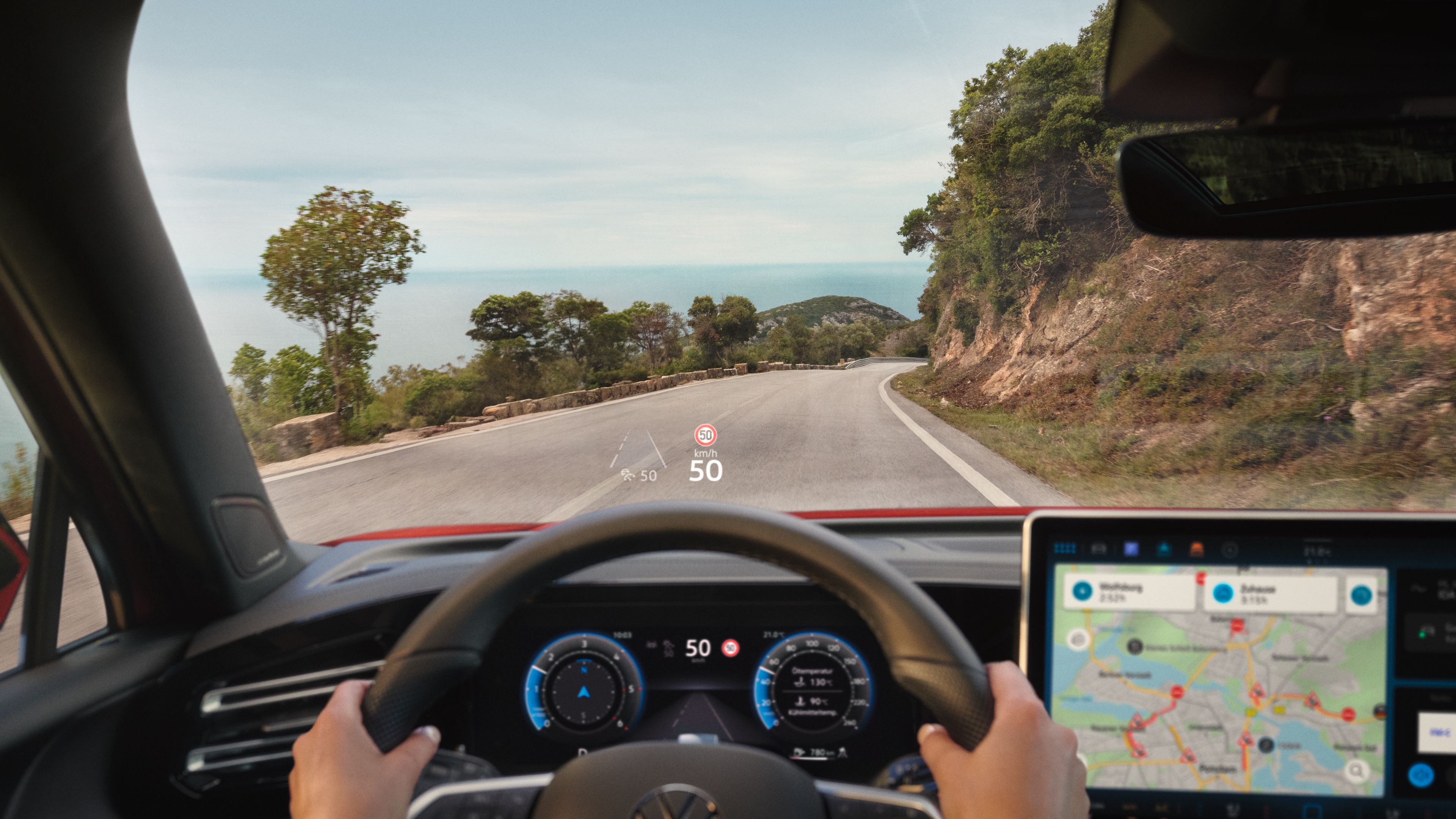 Pohľad do interiéru VW Golf. Za volantom sedí osoba a riadi vozidlo. Vidíme tiež digitálne prístroje, ventiláciu, displej systému infotainment so spustenou navigáciou. Na čelnom skle je zreteľný head-up displej a cez okno vidieť cestu v horách.