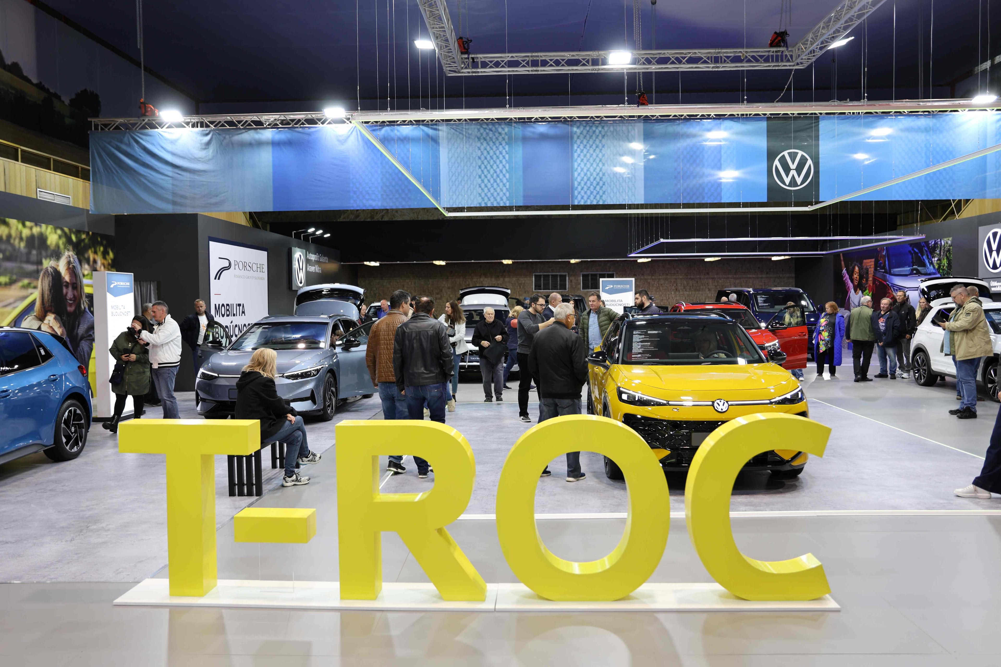 Výstava automobilov Volkswagen, kde je hlavnou atraktivitou žltý model T-Roc. Návštevníci si prezerajú rôzne automobily vystavené v hale. Na stene za autami sú logá Volkswagen a reklamné bannery.