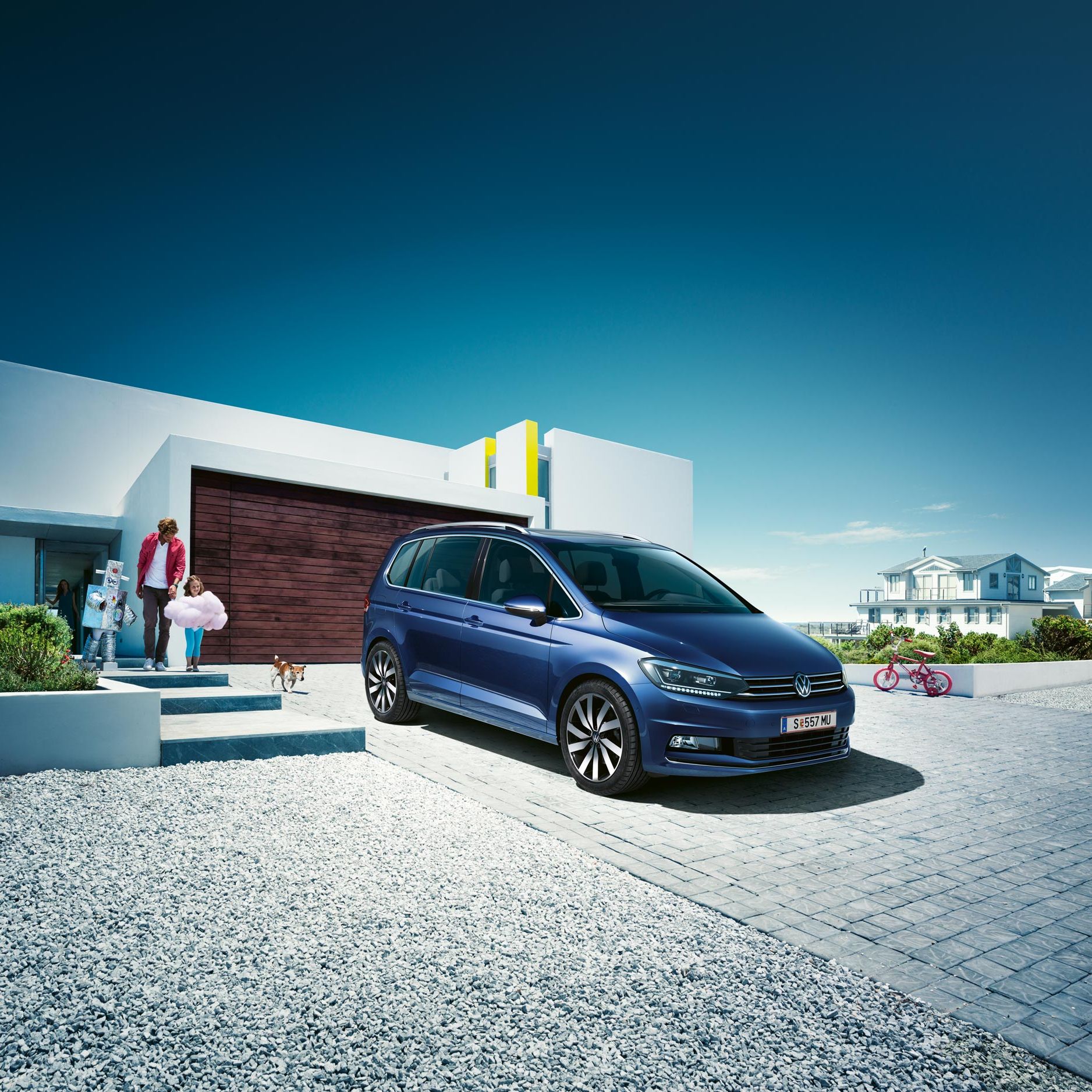 Modrý VW Touran zaparkovaný pred rodinným domom. Z domu vychádza otec s dvomi deťmi, ktoré sú oblečené do kostýmov. Pri nízkom bielom  múriku stojí ružový detský bicykel. V pozadí vidieť ďalšiu veľkú vilu. Obrázku dominuje modrá čistá obloha.