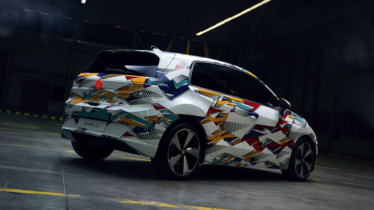 Fotografia zobrazuje auto vo futuristickej hale s vysokým stropom. Vozidlo má moderný dizajn s pestrofarebnou kamuflážou.