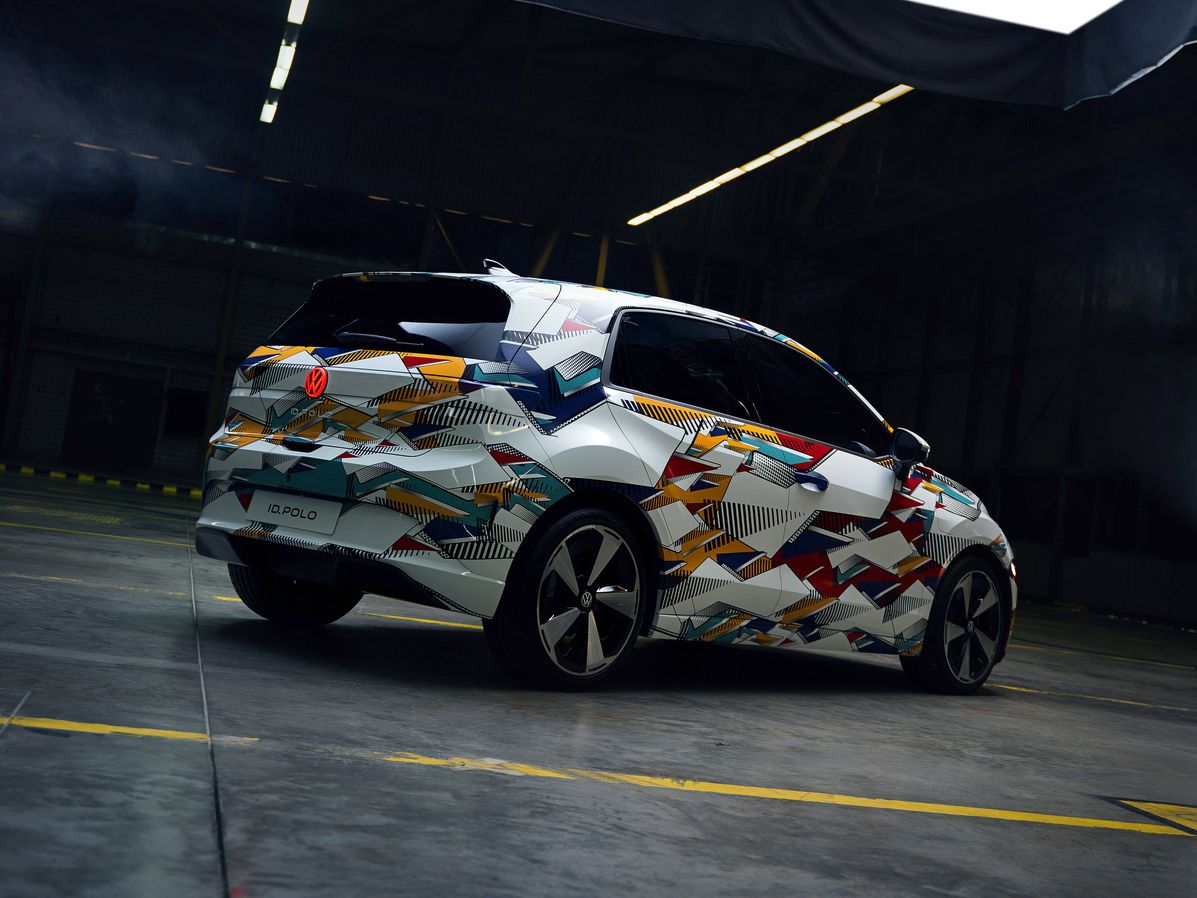 Fotografia zobrazuje auto vo futuristickej hale s vysokým stropom. Vozidlo má moderný dizajn s pestrofarebnou kamuflážou.
