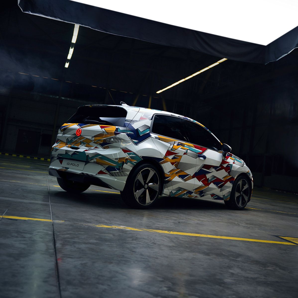 Fotografia zobrazuje auto vo futuristickej hale s vysokým stropom. Vozidlo má moderný dizajn s pestrofarebnou kamuflážou.