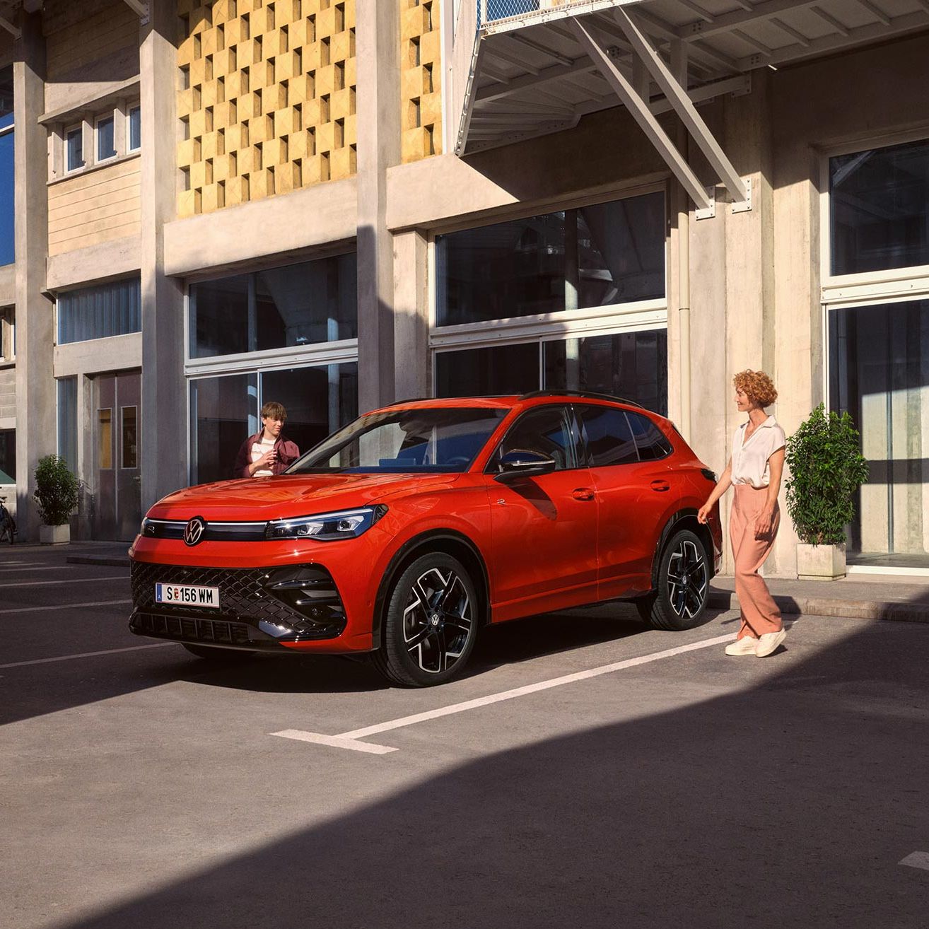 VW Tiguan parkuje pred modernou budovou, približujú sa k nemu dvaja ľudia, žena a mladý muž.