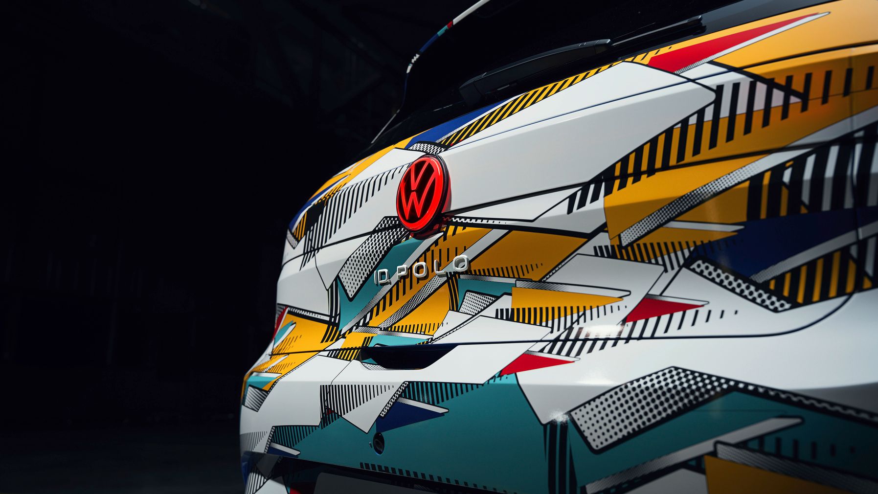 Detailný záber na zadnú časť vozidla Volkswagen s moderným grafickým dizajnom. Zadné svetlo je čiastočne viditeľné a logo VW je červené. Pozadie je tmavé.