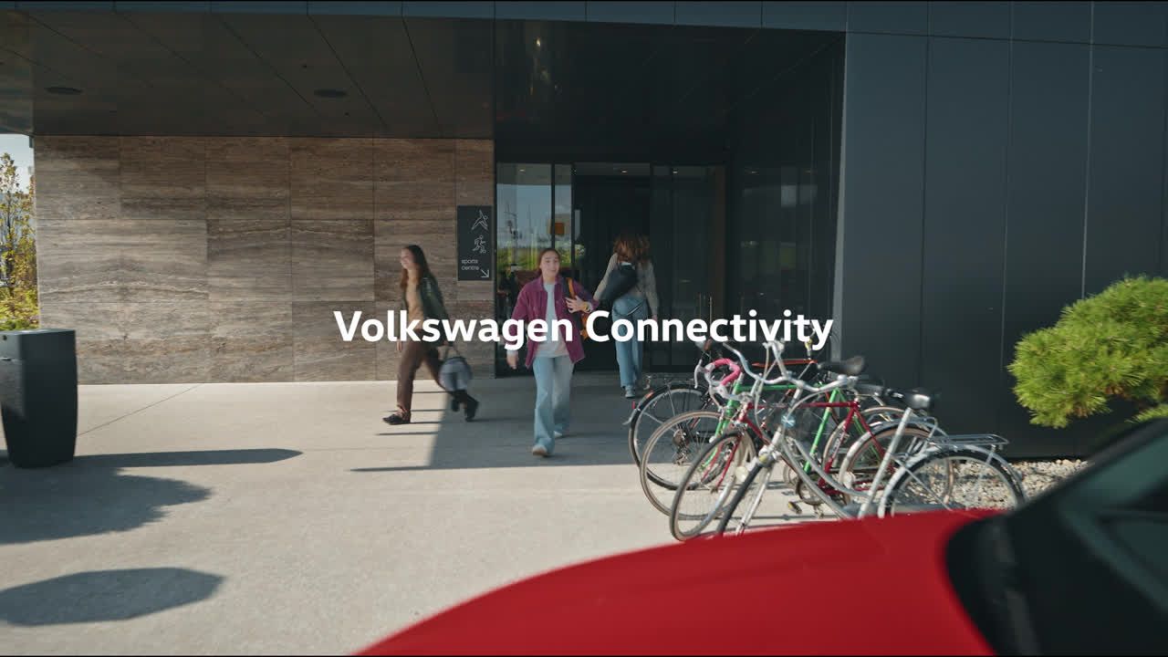Skupina ľudí vychádza z vchodu modernej budovy. Pred budovou je zaparkovaných niekoľko bicyklov. Text na obrázku hovorí "Volkswagen Connectivity".