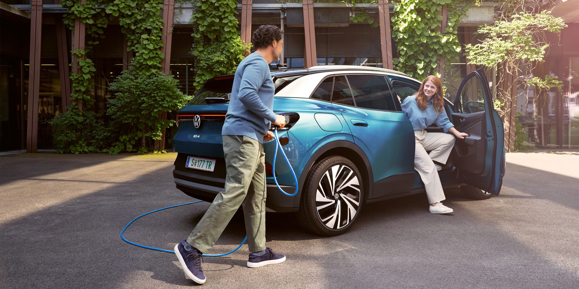 Muž s nabíjacím káblom v ruke kráča k modrému VW ID.4 zaparkovanému na dvore.