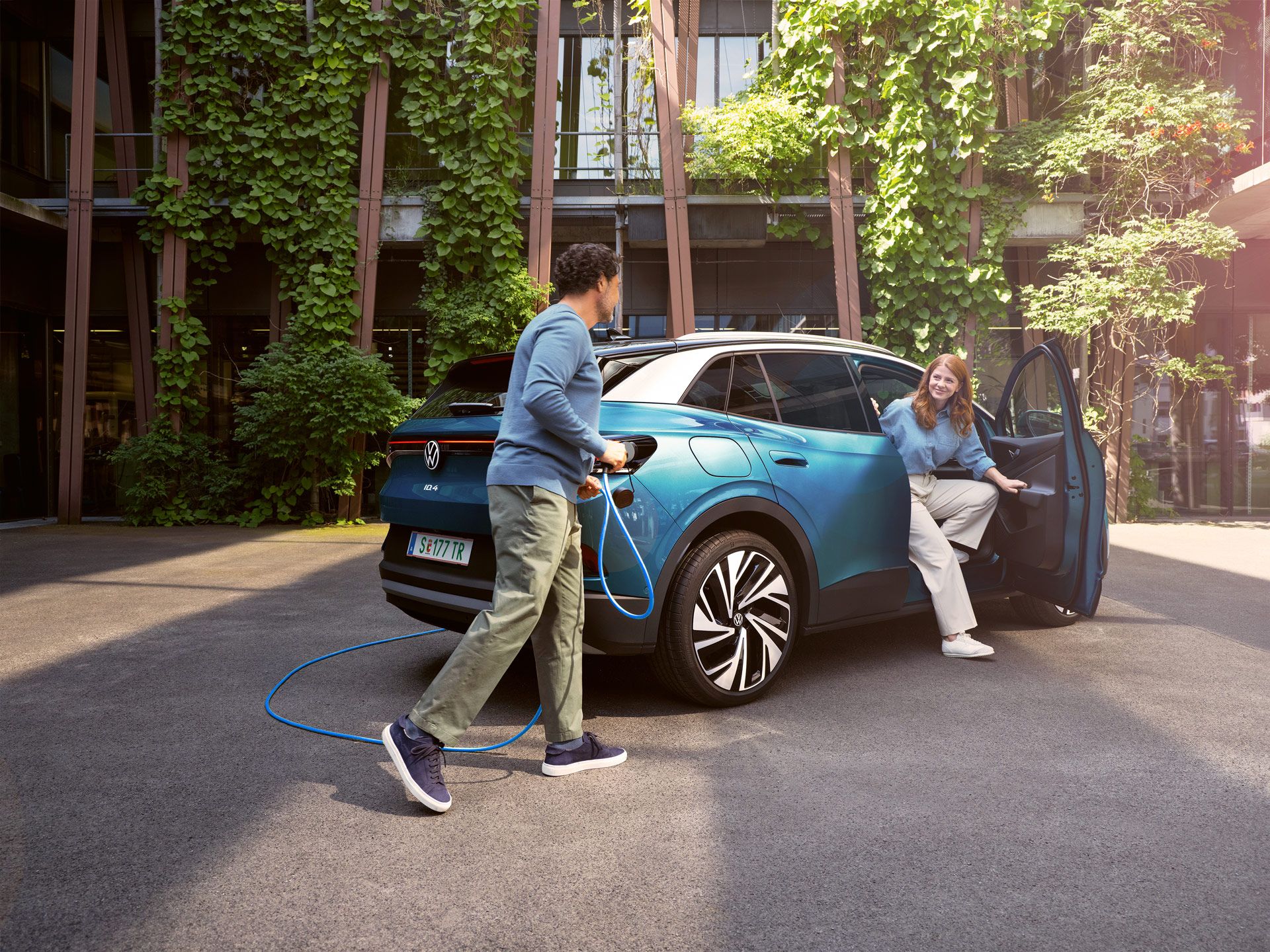 Muž s nabíjacím káblom v ruke kráča k modrému VW ID.4 zaparkovanému na dvore.