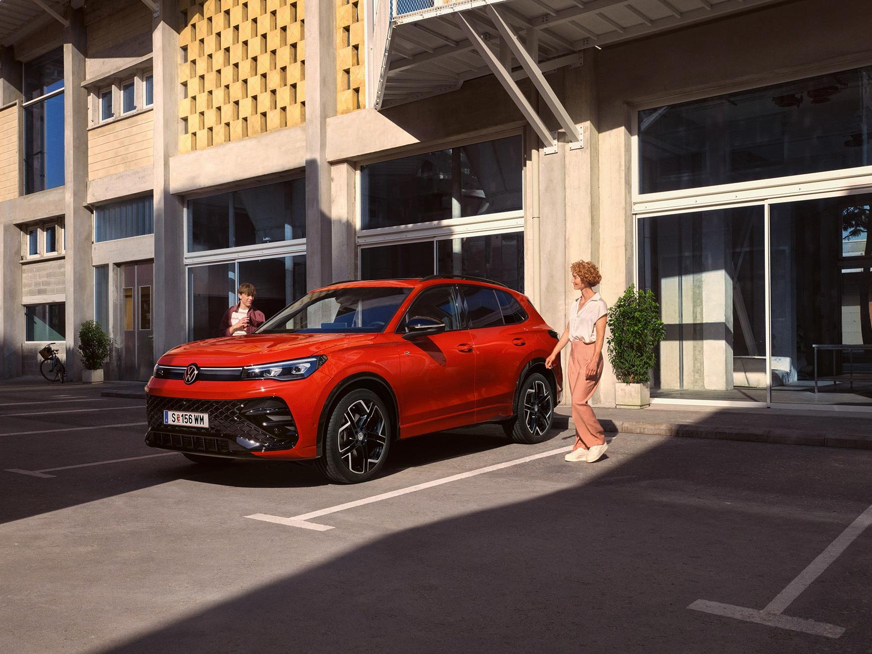 VW Tiguan parkuje pred modernou budovou, približujú sa k nemu dvaja ľudia, žena a mladý muž.