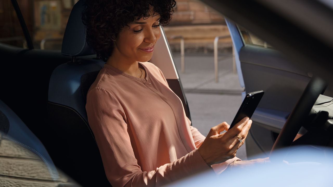 Usmievajúca sa žena na sedadle vodiča zaparkovaného VW T-Cross pozerá na svoj mobilný telefón.